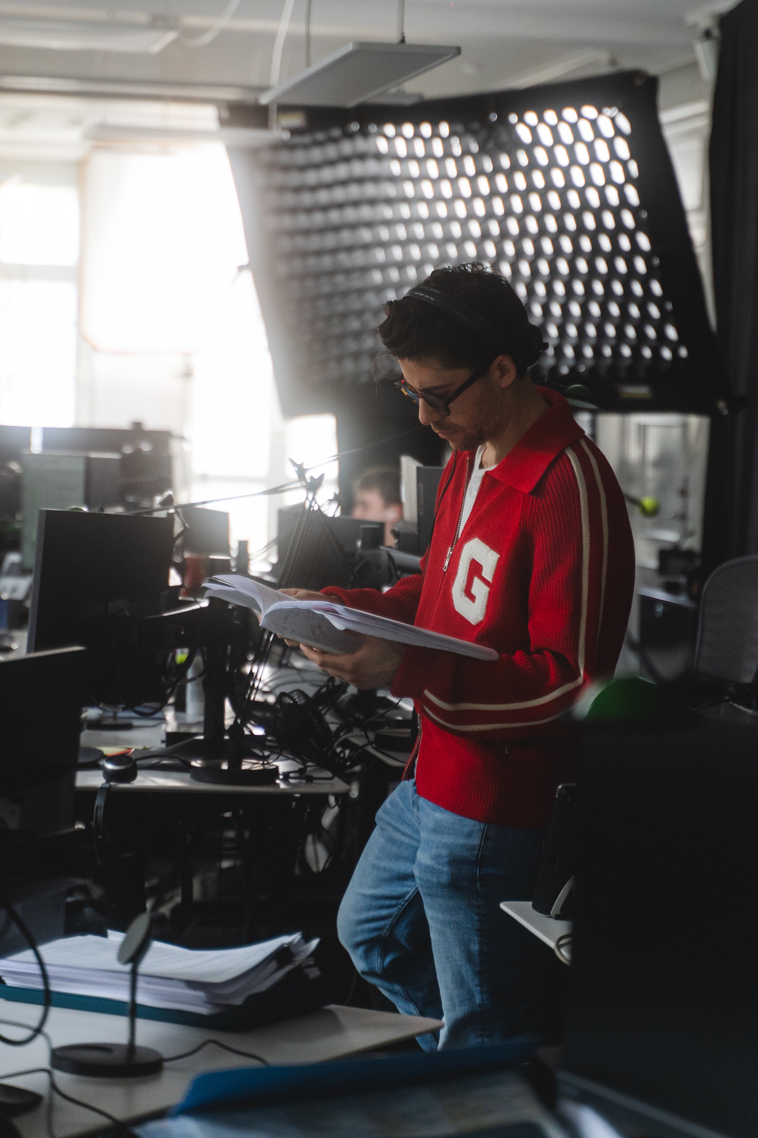A man in a red jacket with a white 'G' on it, glasses, and dark hair, standing in a busy office or studio environment, reading a document or notebook. The workspace has multiple screens, papers, and equipment, with professional lighting and a large w