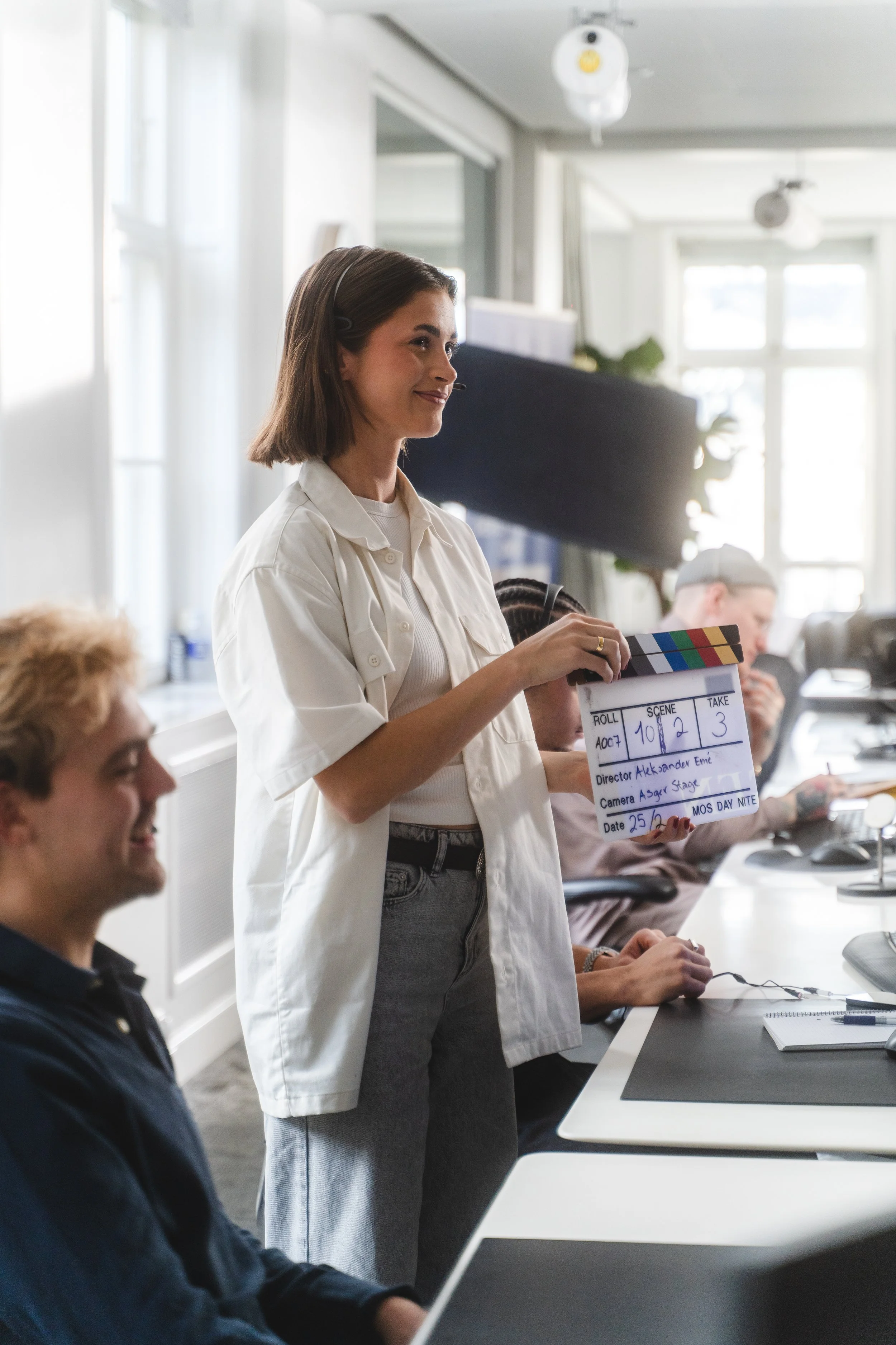 A woman on a film set holding a clapperboard with information, while others work on computers in a bright, modern office.