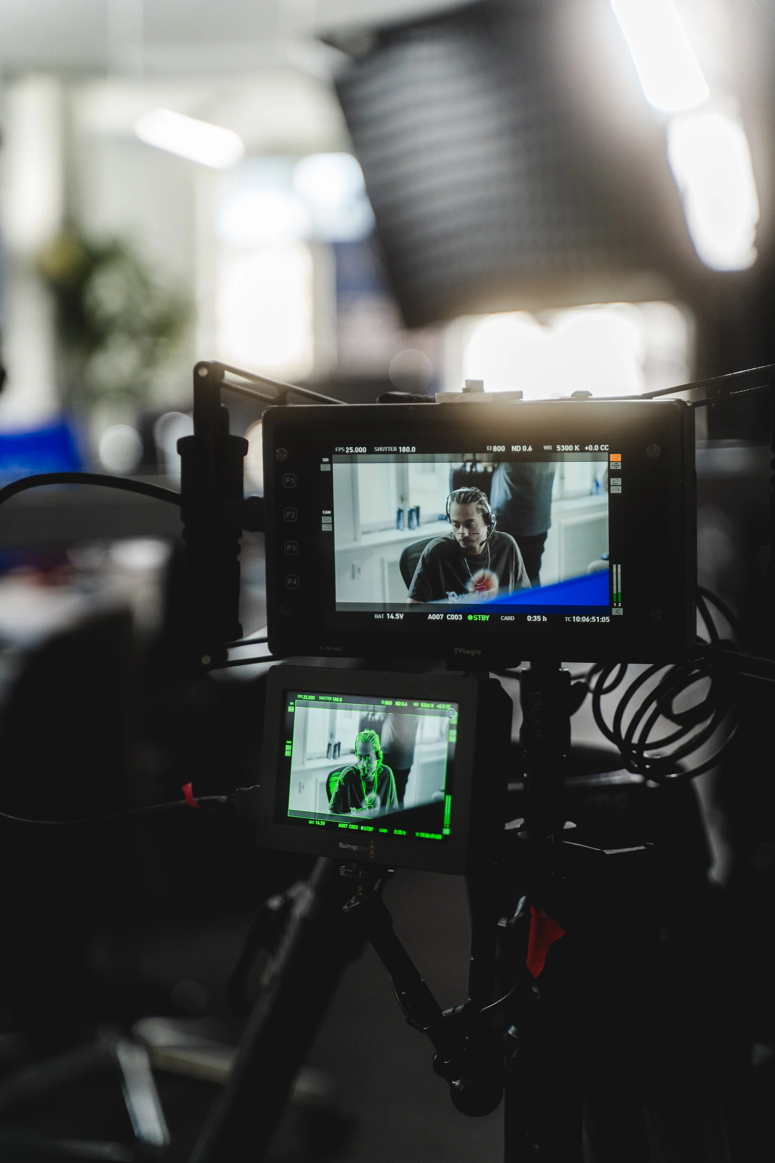 On a film set, two cameras are focused on a woman sitting in a room, with the shot being filmed. The woman is wearing headphones and looking down.