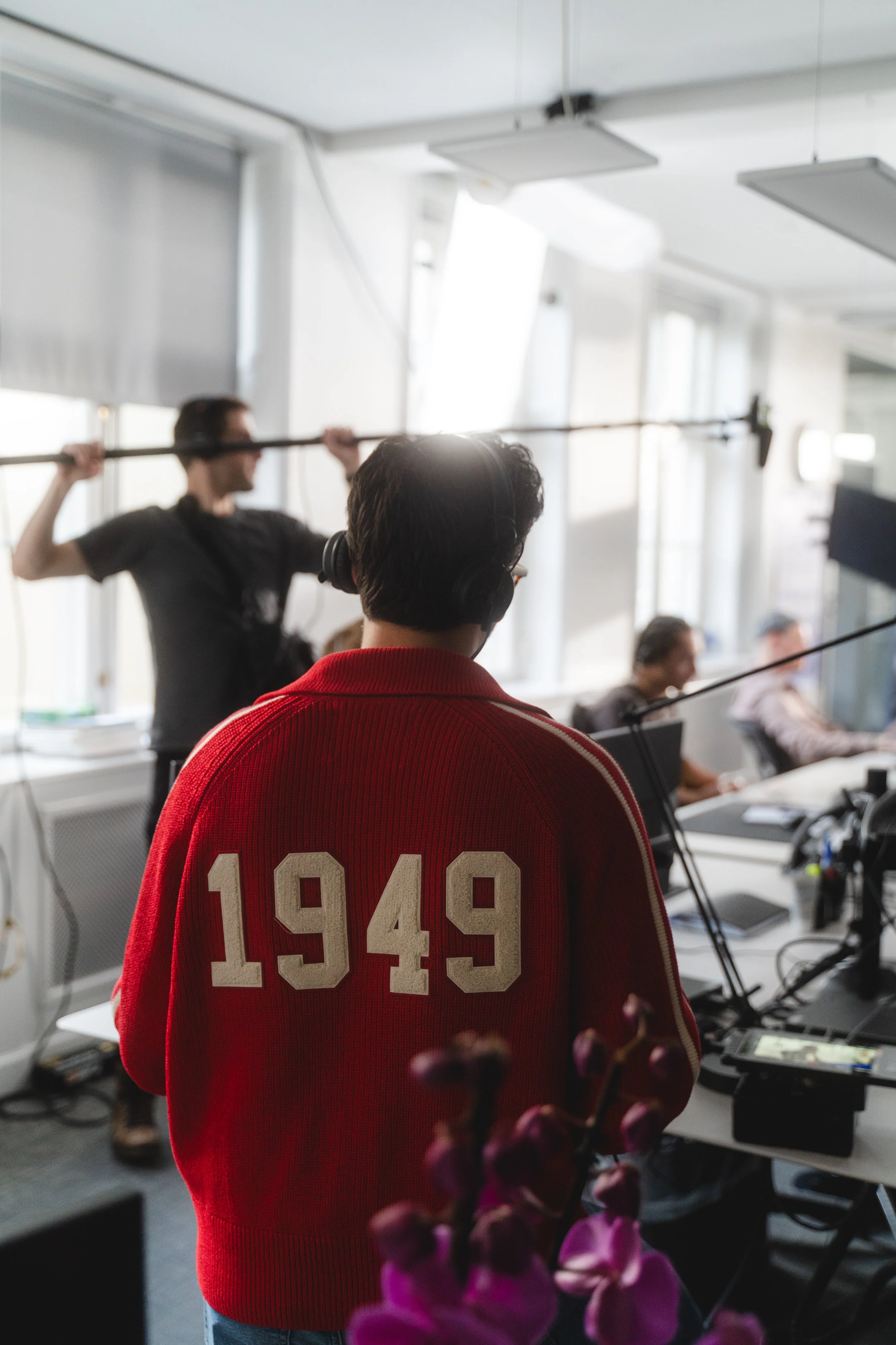 Back view of a person wearing a red jacket with '1949' on the back, in a studio or office setting with people working at desks and a person holding a boom microphone in the background.