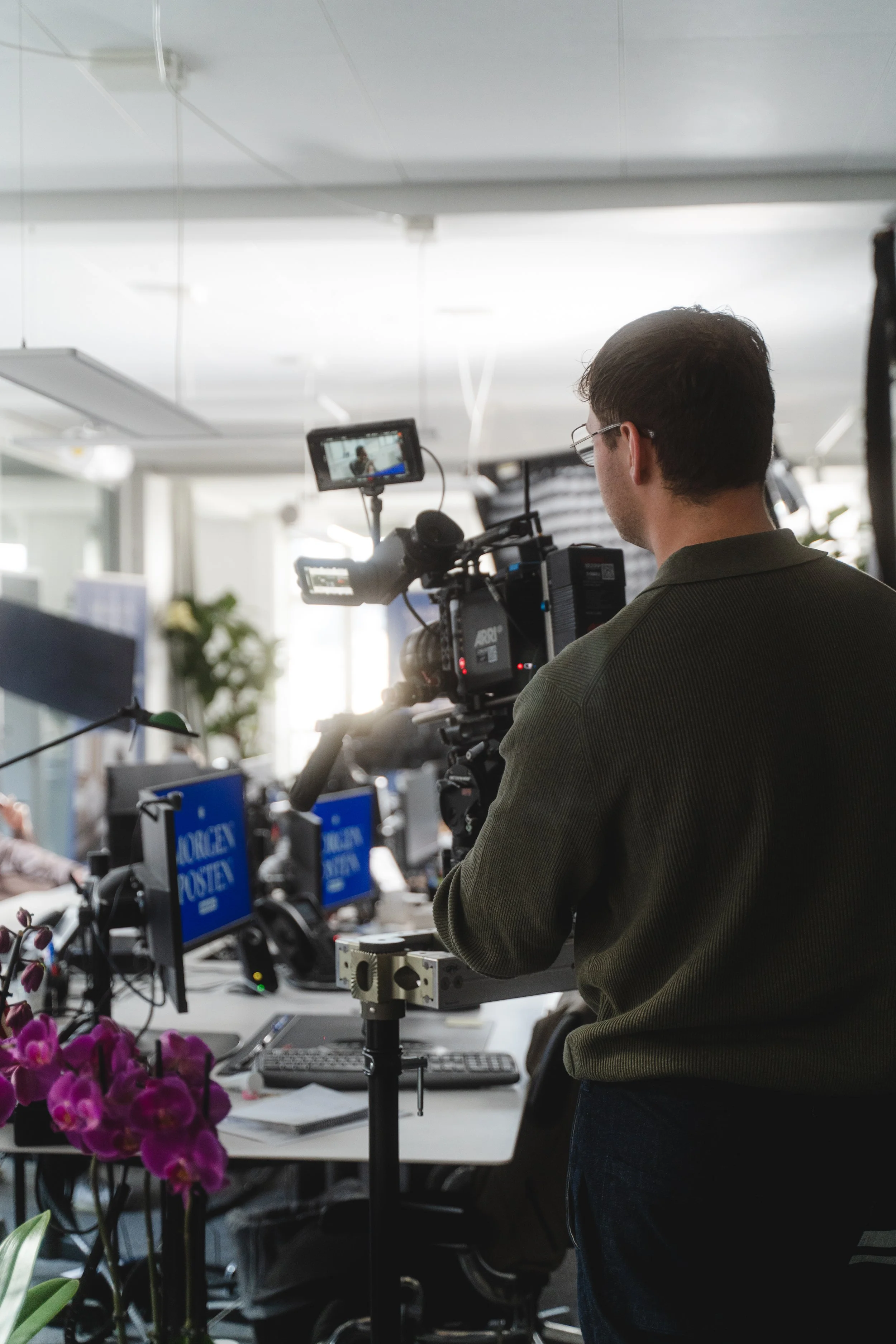 A man operating a professional video camera in an office, with multiple monitors and office equipment visible in the background.