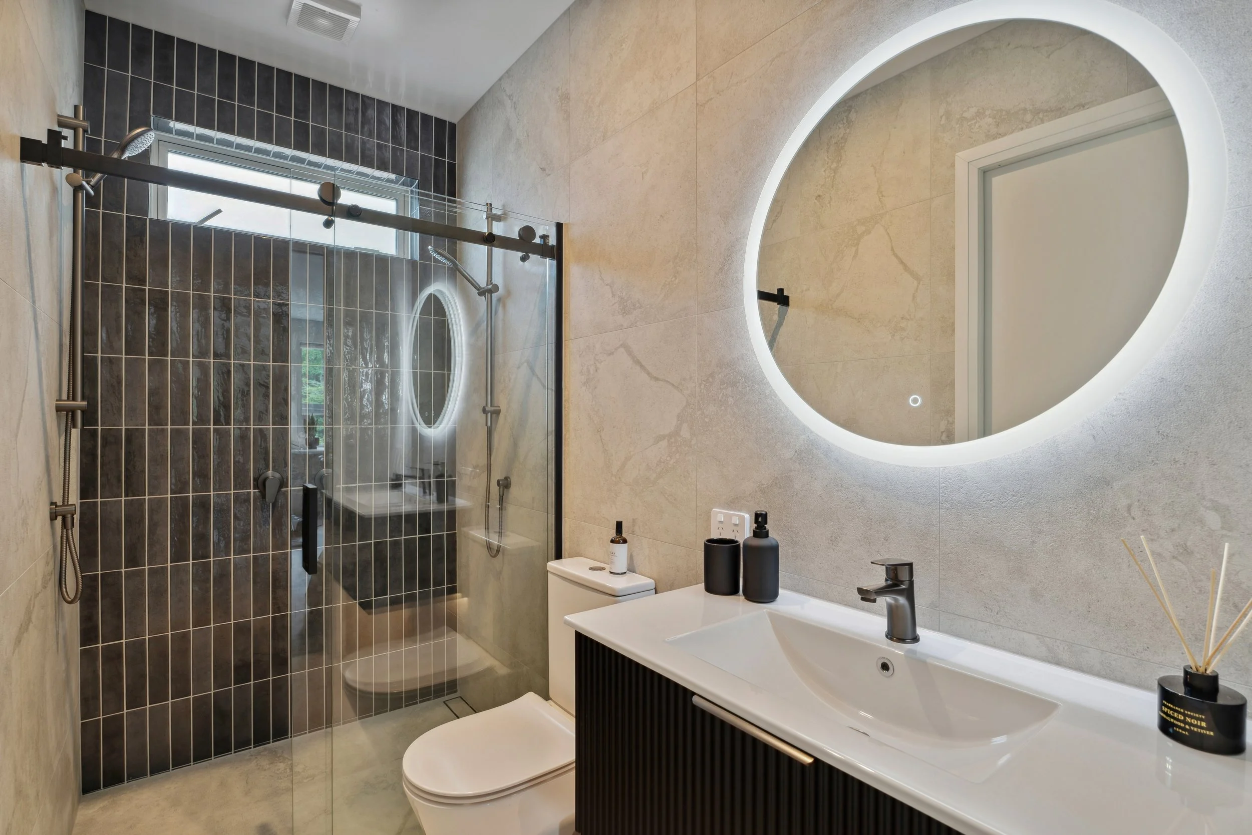 Modern bathroom with walk-in shower enclosed in glass, large oval backlit mirror, beige textured walls, and a black and white vanity with toiletries.