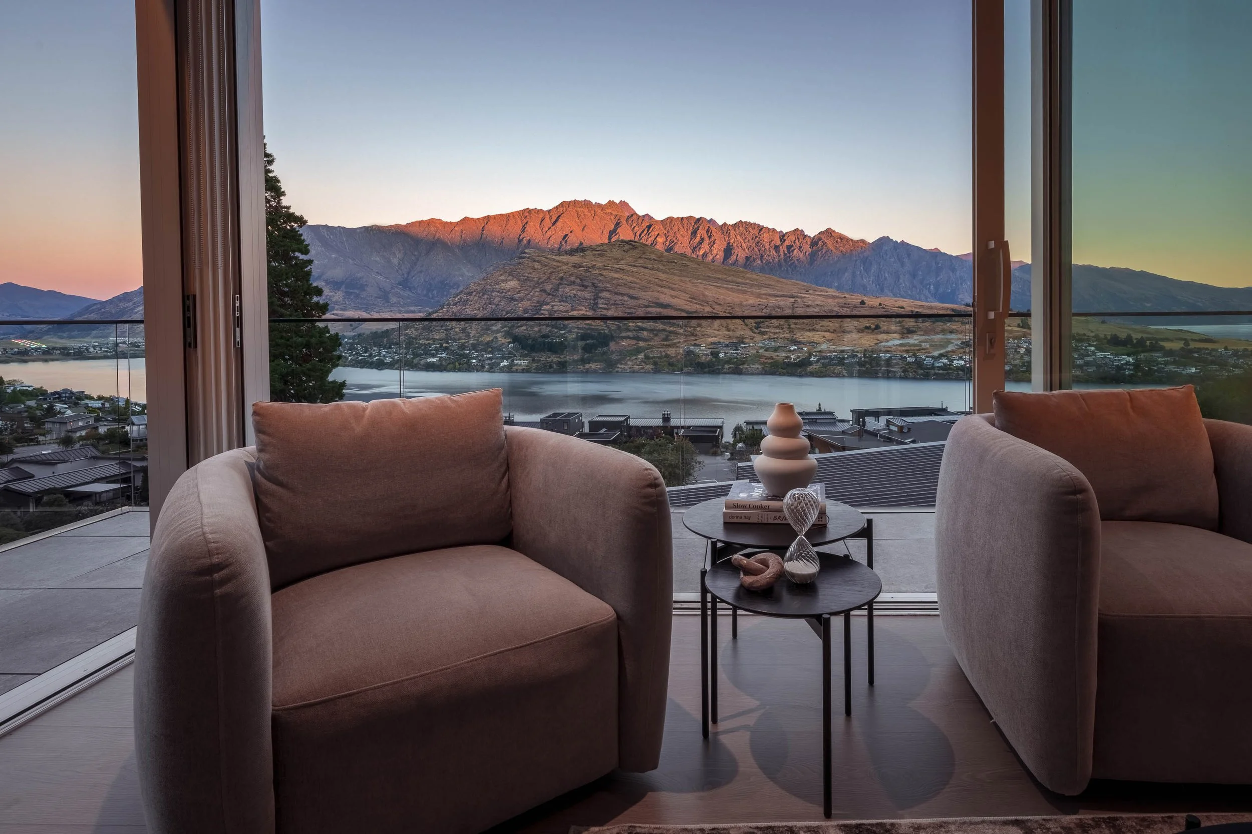 Living room with two beige armchairs and a small side table with decorative vases, overlooking a river and mountain landscape at sunset through large glass doors.