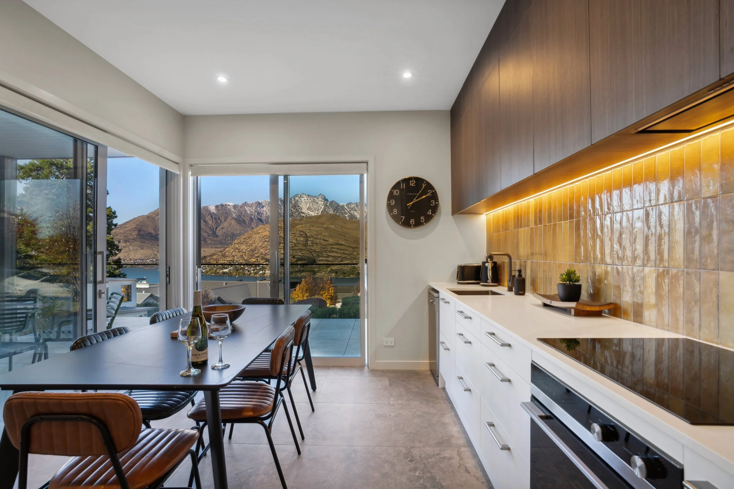 Modern kitchen and dining area with mountain view through large glass sliding doors, featuring a black wall clock, white cabinets, a black stove, and a dining table set for wine and food.