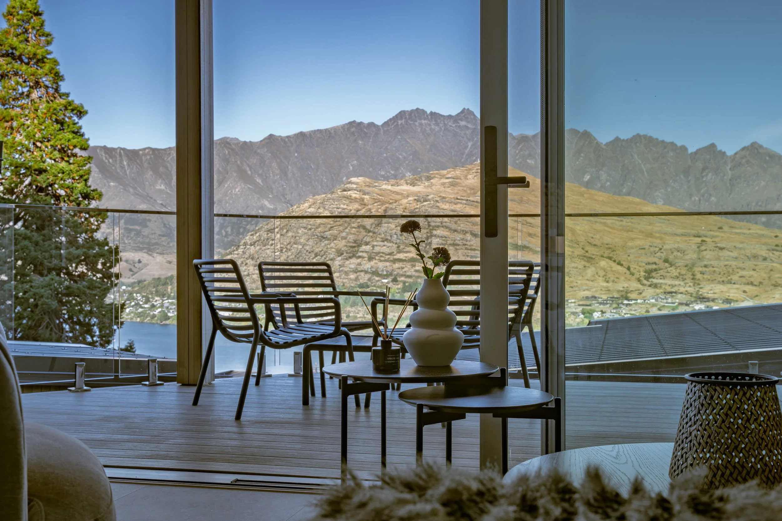Interior living space with floor-to-ceiling glass sliding doors leading to a deck with outdoor chairs, overlooking mountains, a lake, and a hillside.