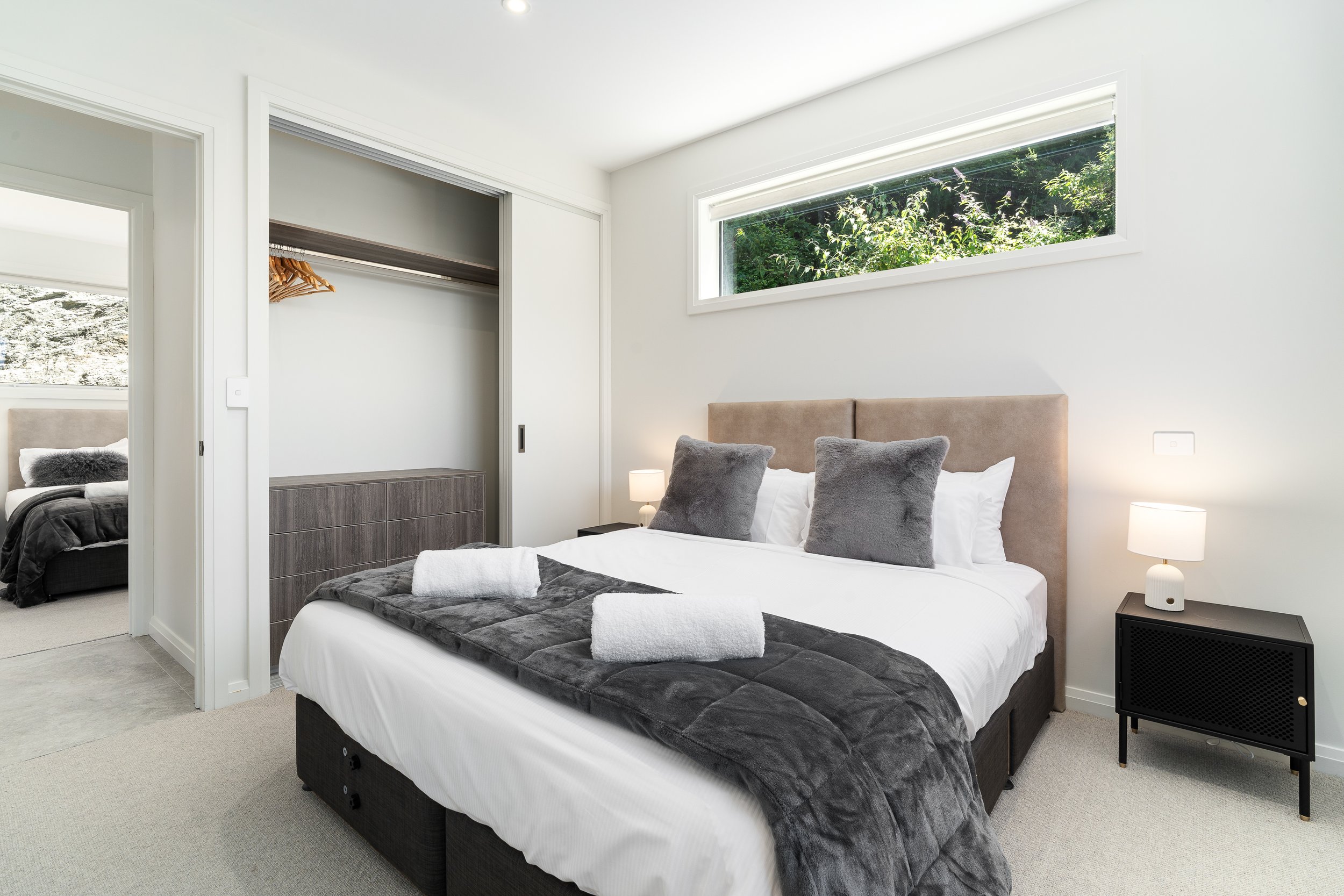 Modern bedroom with a bed, gray pillows, white and gray bedding, and a black nightstand with a lamp, natural light from a horizontal window, and a closet in the background.