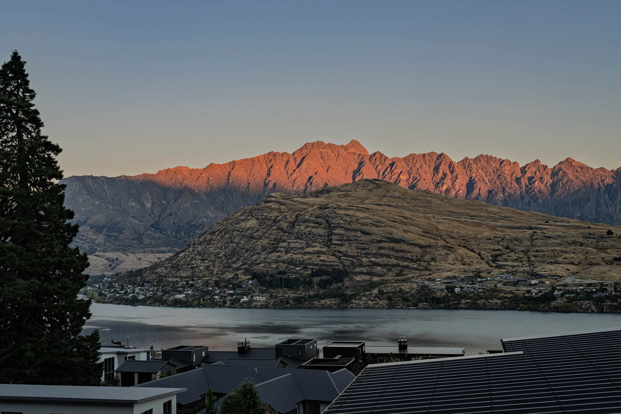 Scenic view of a mountain range at sunset with houses in the foreground and a body of water reflecting the sky.