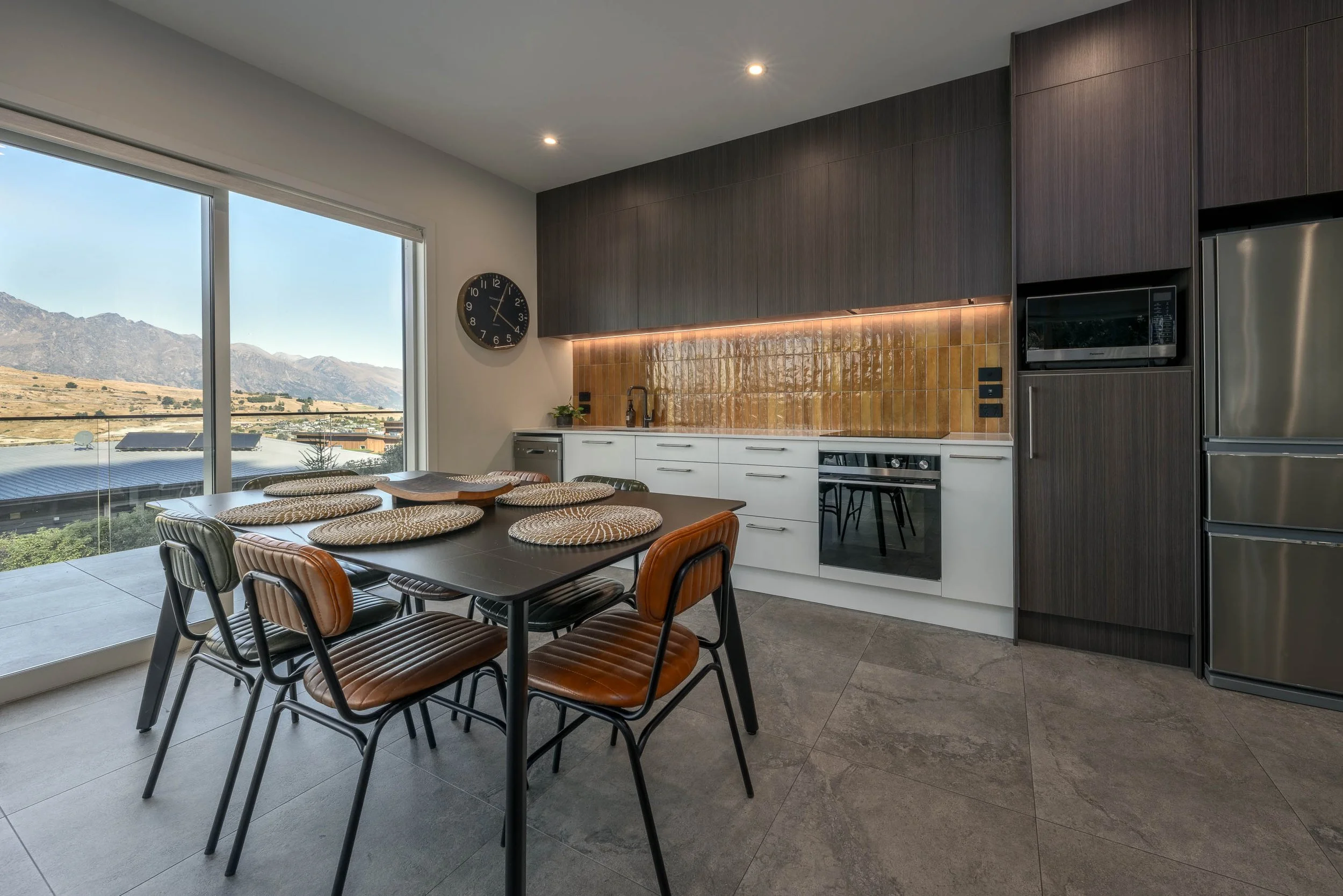 Modern kitchen with white lower cabinets, dark upper cabinets, a yellow tile backsplash, a black clock on the wall, stainless steel appliances, a black dining table with six brown chairs, and a large sliding glass door revealing a mountain landscape 