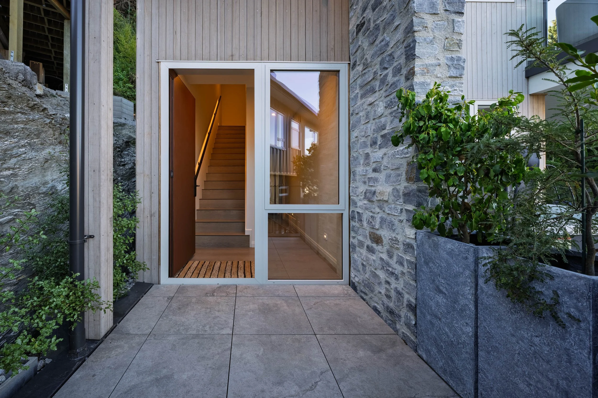 Modern exterior entrance to a house featuring a glass sliding door, concrete tiles on the porch, a stone accent wall, and a planter with green foliage. Inside, a staircase illuminated by warm lighting is visible.