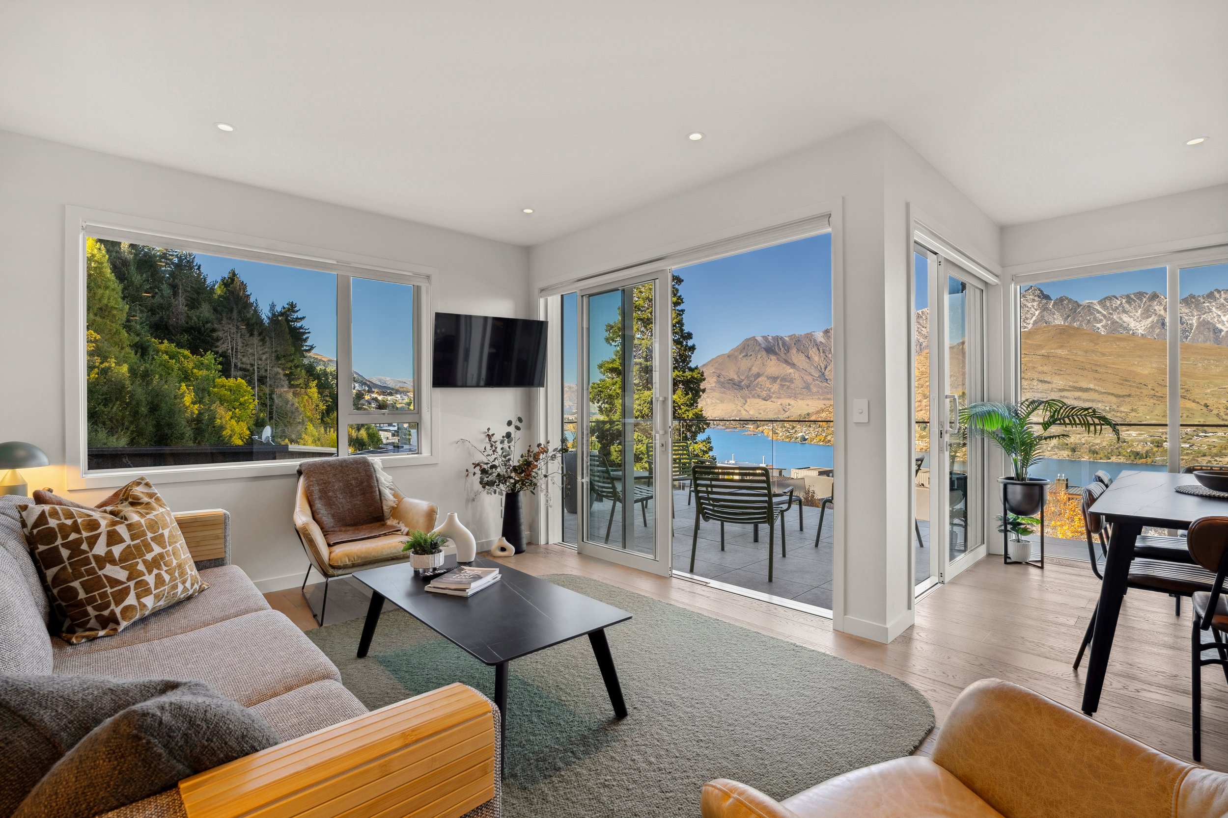 Living room with large windows and sliding glass door overlooking mountain and lake view, furnished with a sofa, armchair, coffee table, television, and dining table with chairs.