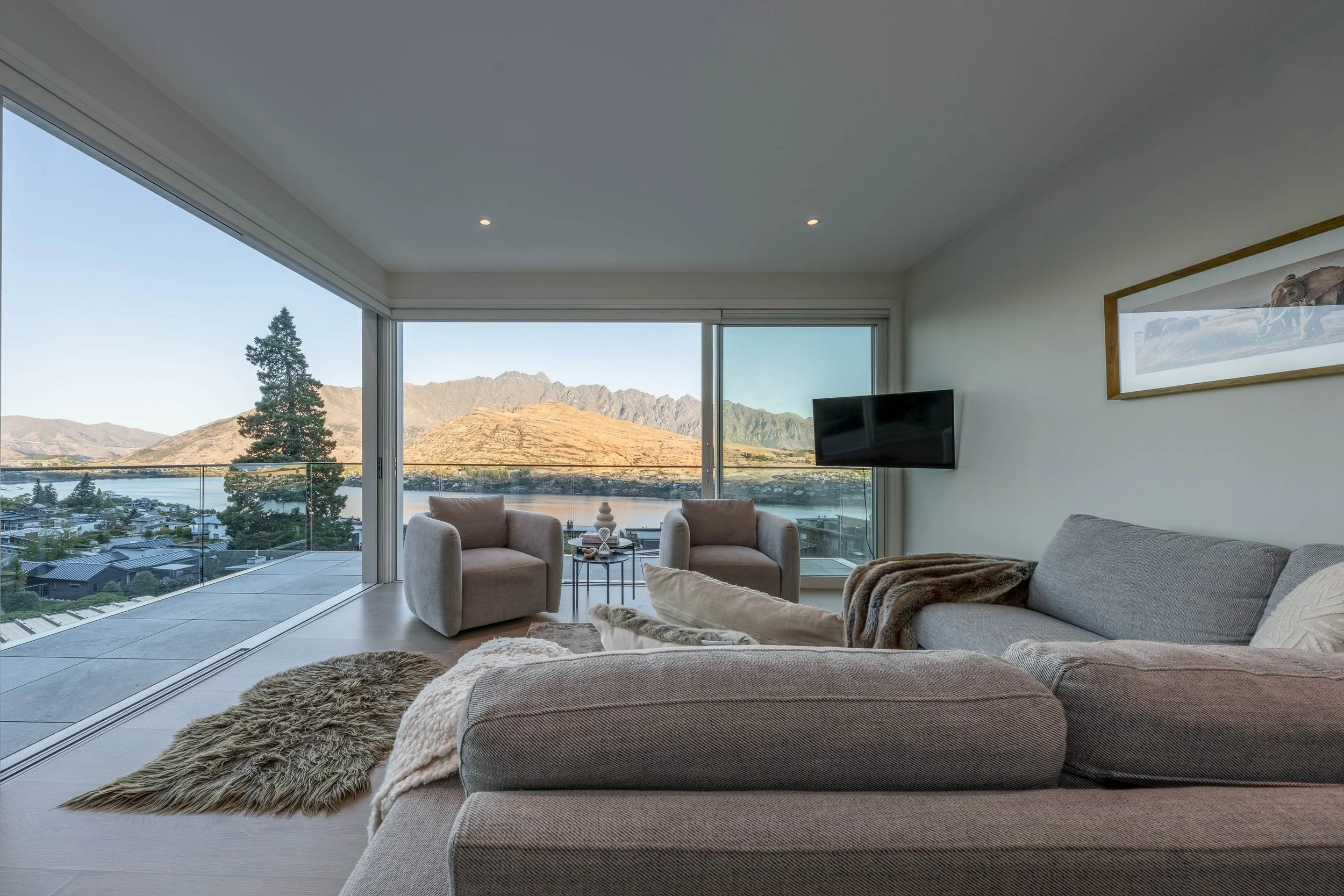 Living room with large windows overlooking mountains and lake, beige sofa with throw blankets, two armchairs, a small side table with decorative objects, wall-mounted TV, framed landscape art, and a balcony with glass railing.