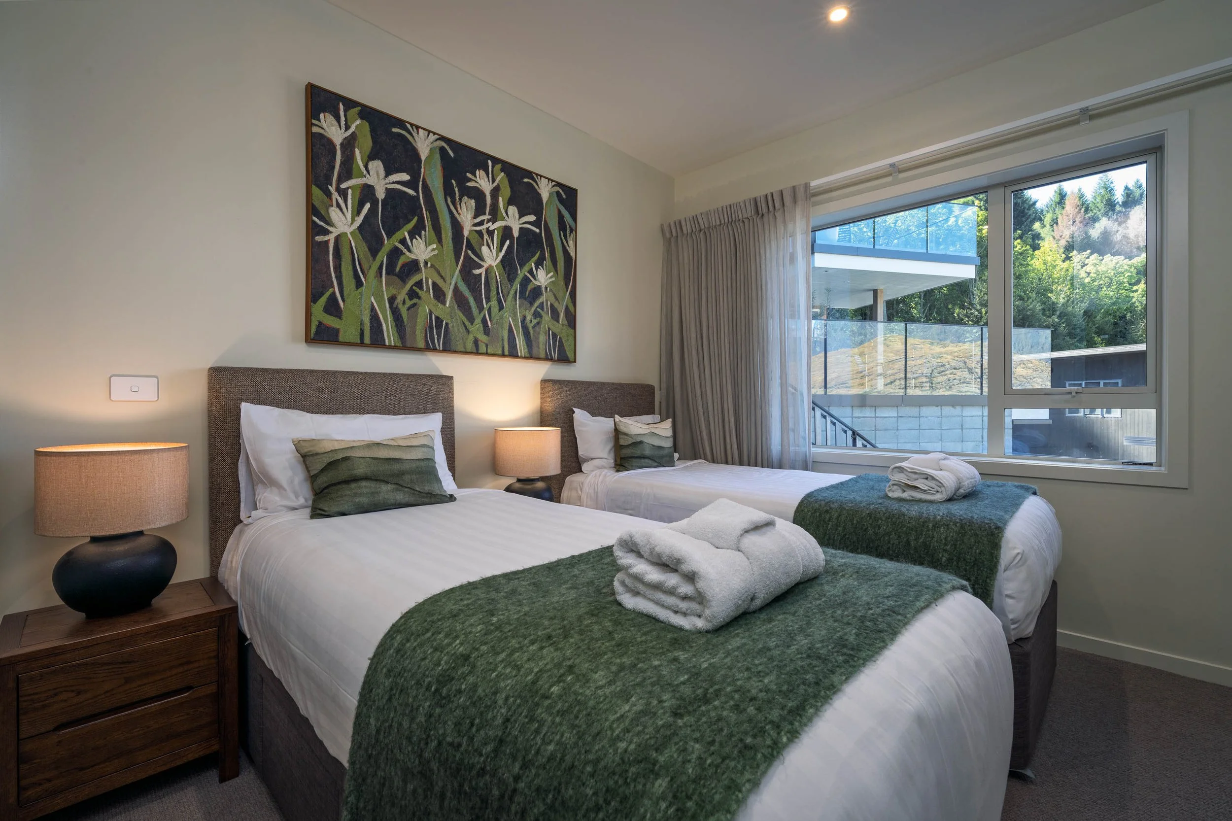 A bedroom with two beds, each with green blankets and rolled white towels, nightstands with lamps, a large window showing an outdoor view, and a botanical painting above the beds.
