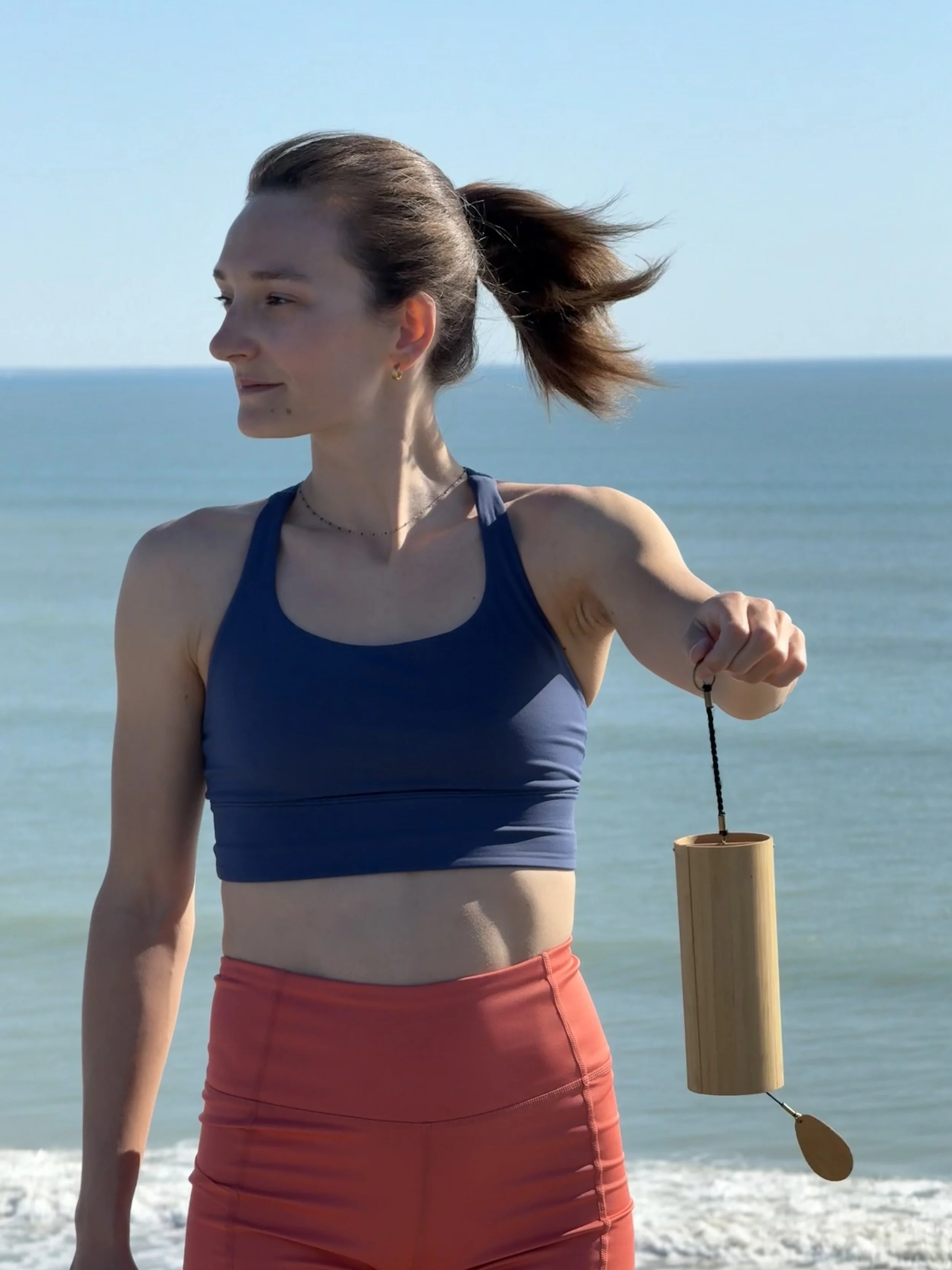 Jeune femme en tenue de sport sur la plage, tenant une lanterne en bambou suspendue par une corde, avec la mer en arrière-plan.