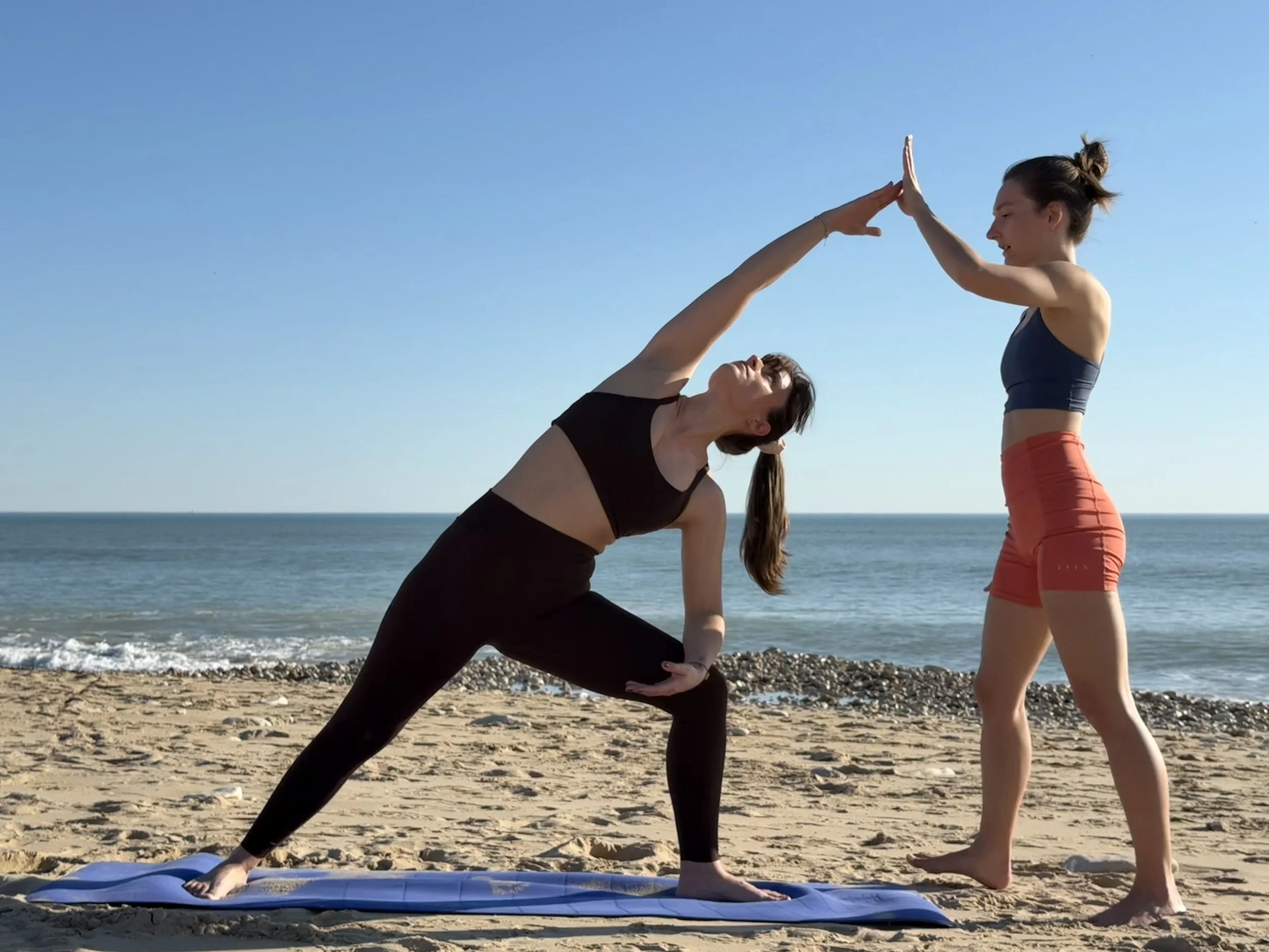 Deux femmes pratiquent le yoga sur la plage, une en position de guerrier et l'autre en position de prière, sous un ciel clair avec la mer en arrière-plan.