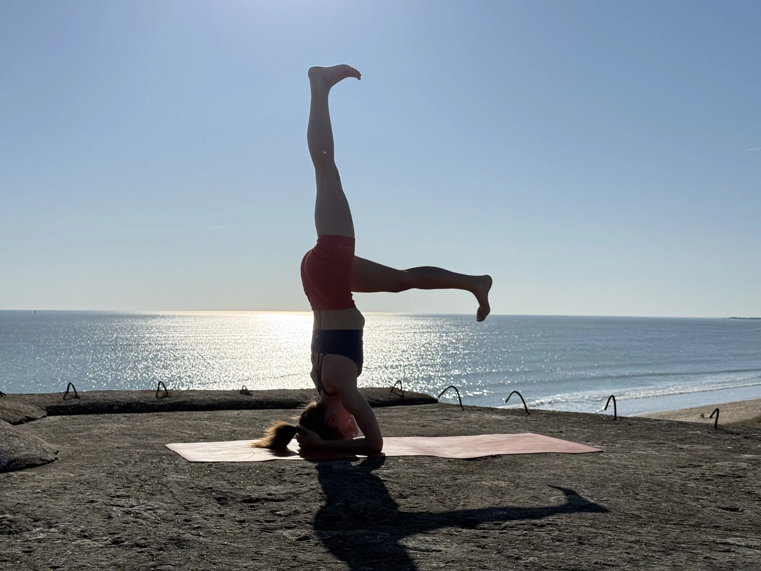Une personne réalise une posture de yoga en équilibre sur la tête, avec les jambes en position de poirier, au bord de la mer lors d'un coucher de soleil.