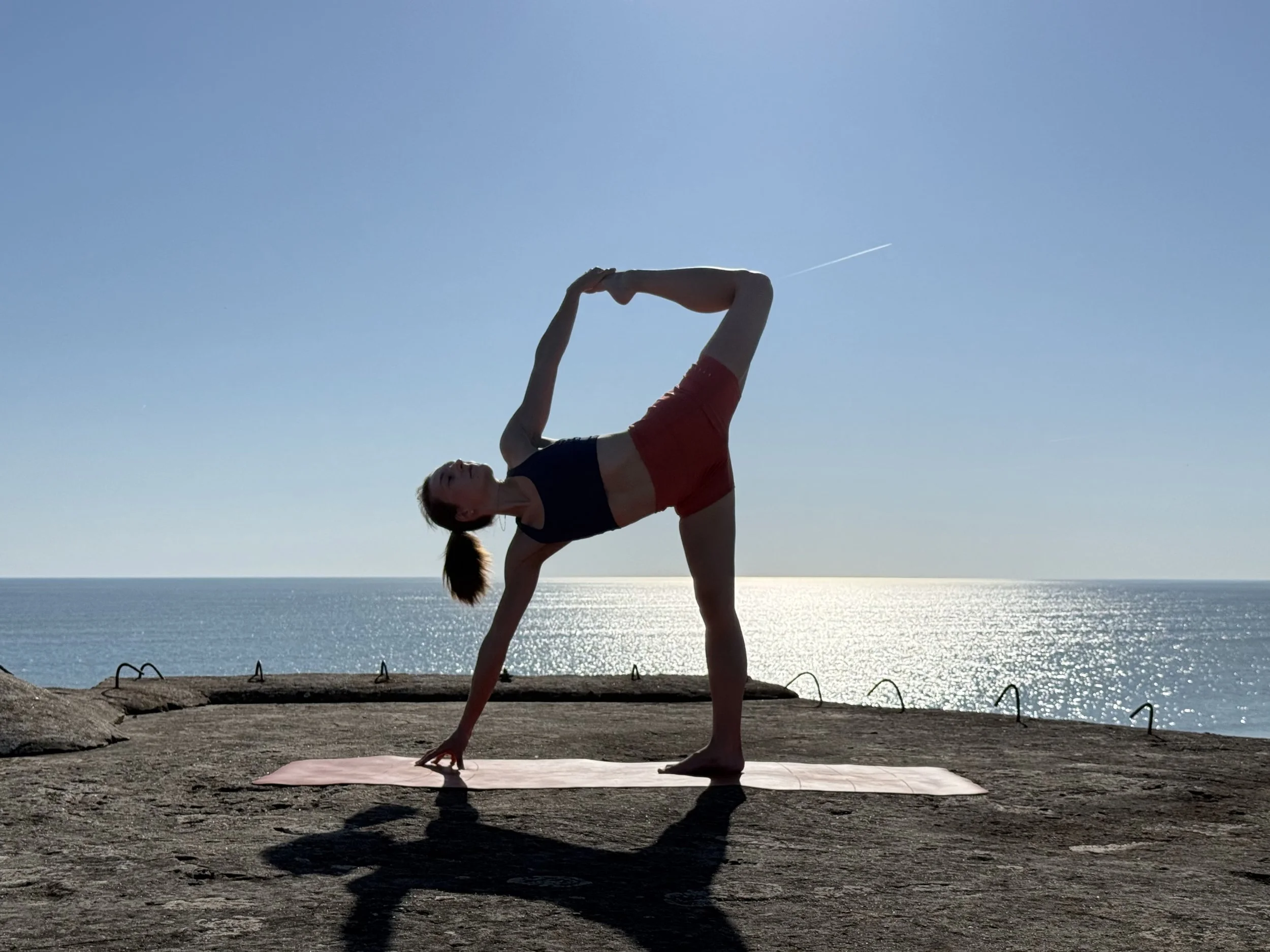 Une femme pratique le yoga en équilibre sur une main sur un tapis près de la mer, avec un ciel clair en toile de fond.