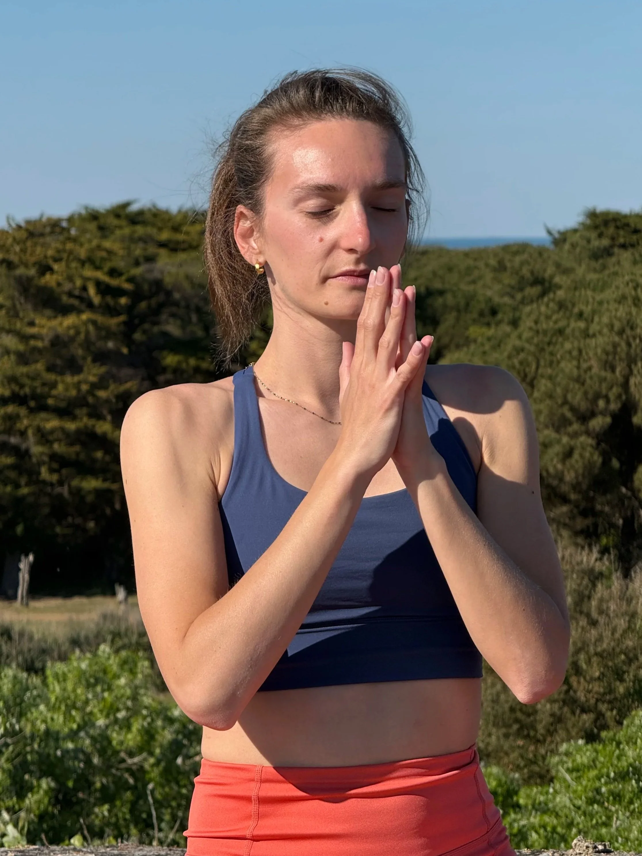 Une femme pratique la méditation et le yoga en plein air, avec les mains jointes devant le visage, les yeux fermés, dans un environnement naturel avec des arbres et un ciel bleu.