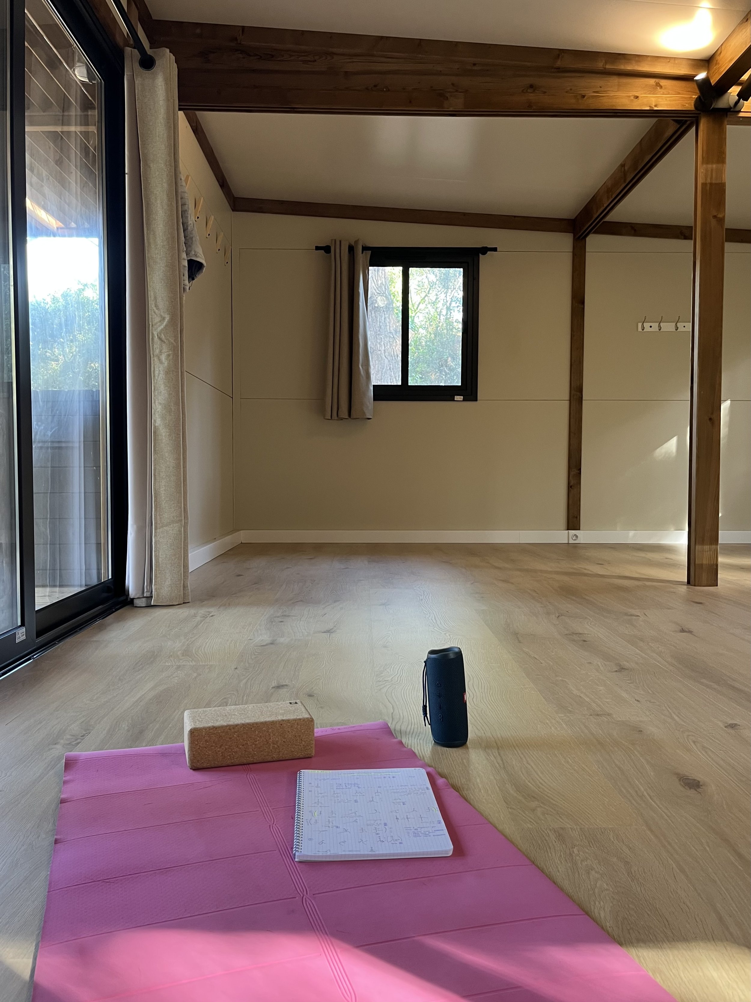 Une salle de yoga ou méditation avec un tapis rose, un bloc en liège, un carnet avec des notes, une bouteille d'eau ou haut-parleur, dans une pièce lumineuse avec de grandes fenêtres, des murs en bois et un sol en bois.
