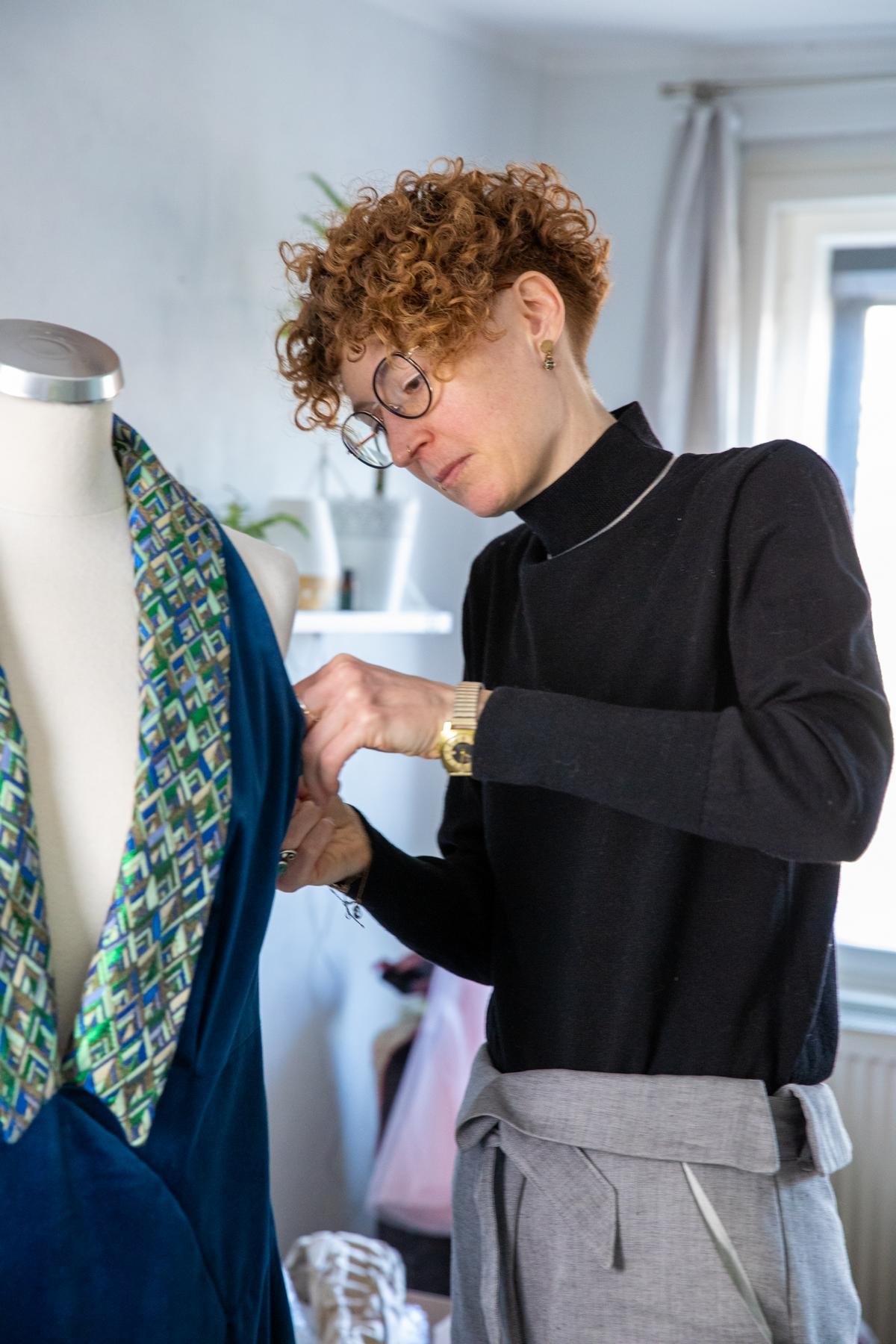 A woman with curly red hair, glasses, and earrings working on a garment on a mannequin, in a well-lit room.