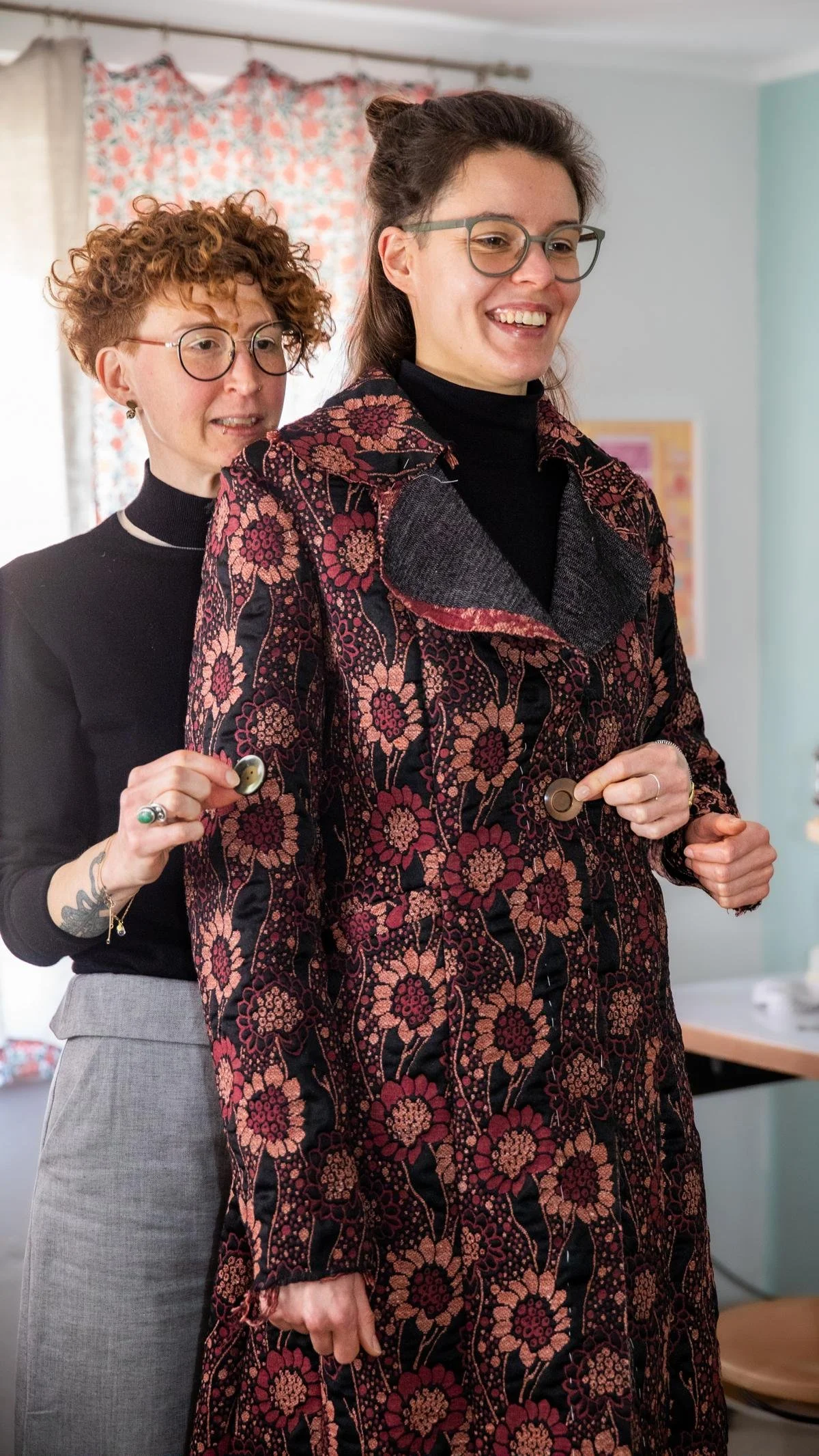 Two women, one in a floral patterned coat and glasses and another with curly red hair, glasses, and tattoos, standing behind and adjusting the other's buttoned coat.