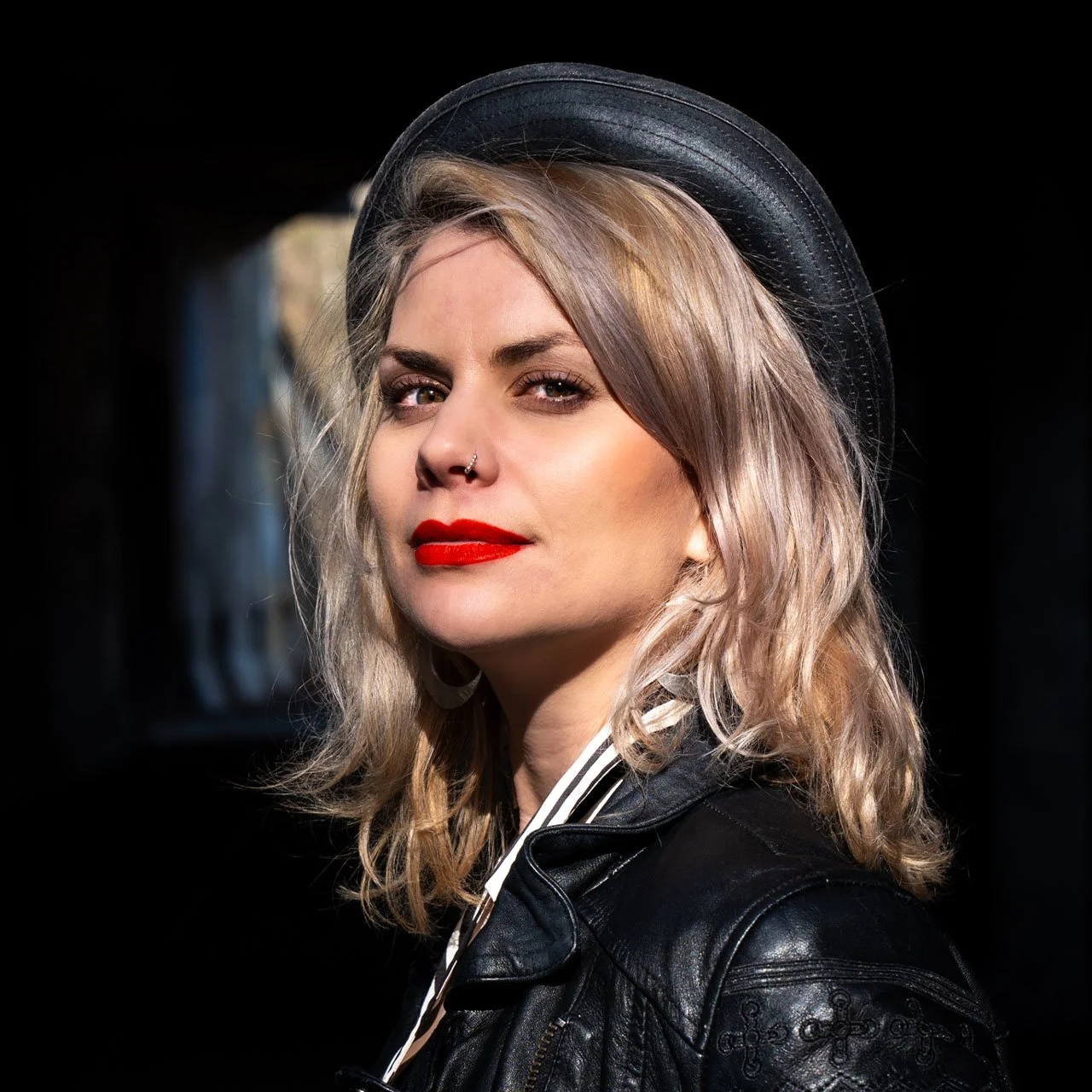 A woman with wavy blonde hair, red lipstick, and a nose piercing, wearing a black hat and leather jacket, looking at the camera against a dark background.