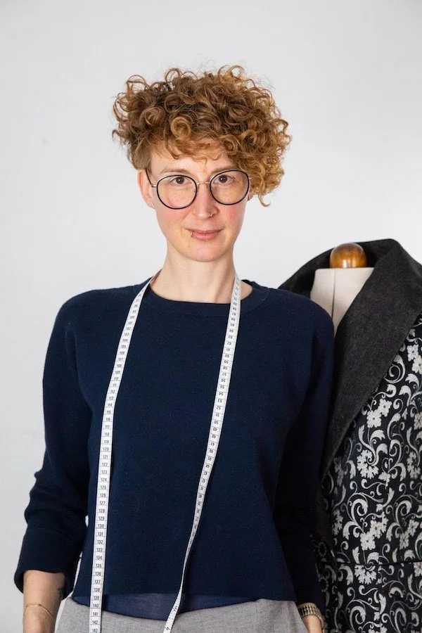 A woman with curly short hair, wearing glasses and a dark blue sweater, stands next to a mannequin dressed in a black and gray patterned blazer with a measuring tape around her neck.