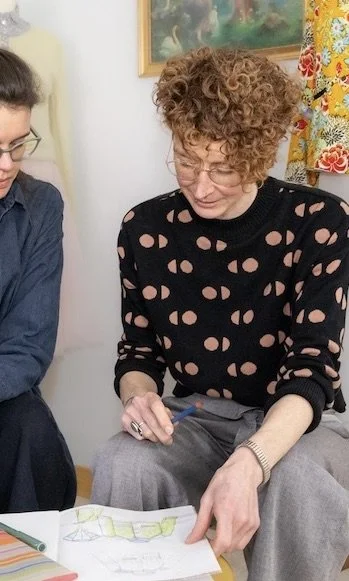 Two women sit together, looking at a sketch or drawing on a piece of paper, in a room with colorful curtain and artwork in the background.