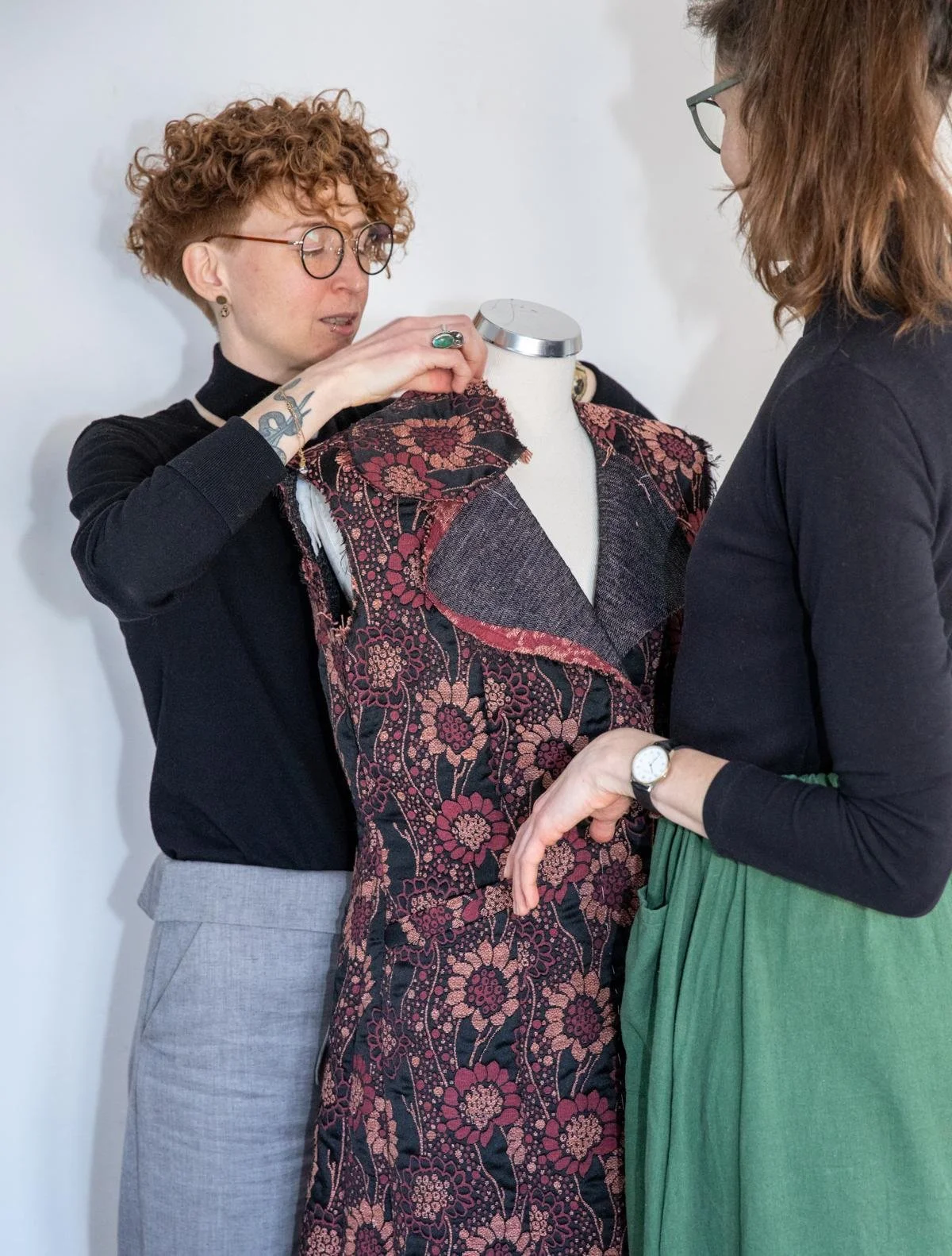Two women are examining a floral patterned fabric on a dress form. One woman with curly hair and glasses is adjusting the fabric, while the other woman is observing.