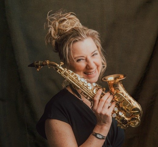 Woman with blonde hair in a bun smiling and holding a gold saxophone.