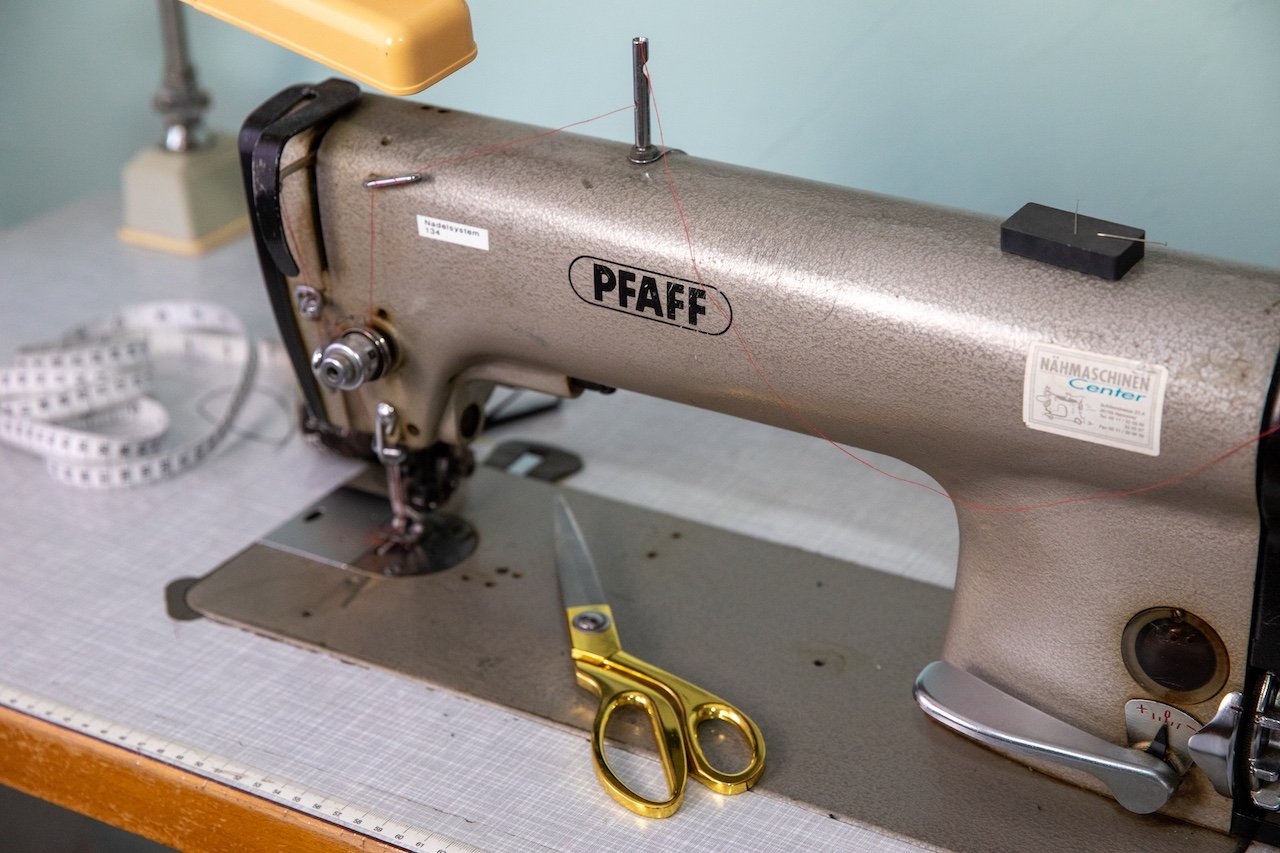 A vintage beige Pfaff sewing machine on a work table with gold-handled scissors, a folded measuring tape, and a yellow lamp in the background.