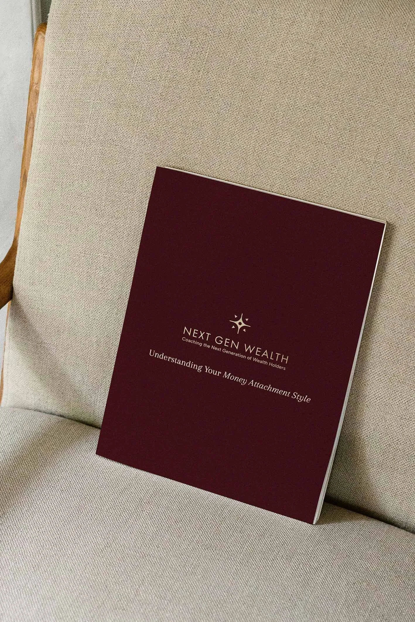 A burgundy-colored booklet titled "Next Gen Wealth" resting on a beige upholstered chair. The subtitle reads "Coaching the Next Generation of Wealth Holders" and the text says "Understanding Your Money Attachment Style."