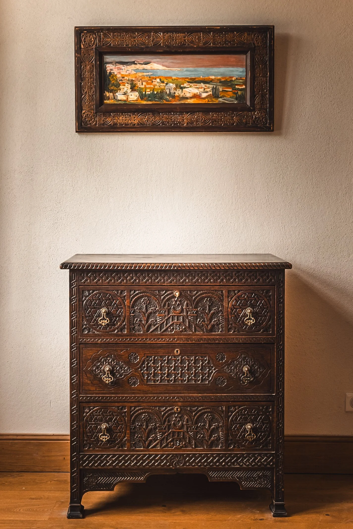 A dark carved wooden chest with multiple drawers and a carved top, in front of a white wall. Above it, a landscape painting with a dark carved wooden frame depicts a rural scene with a sky, buildings, and trees.