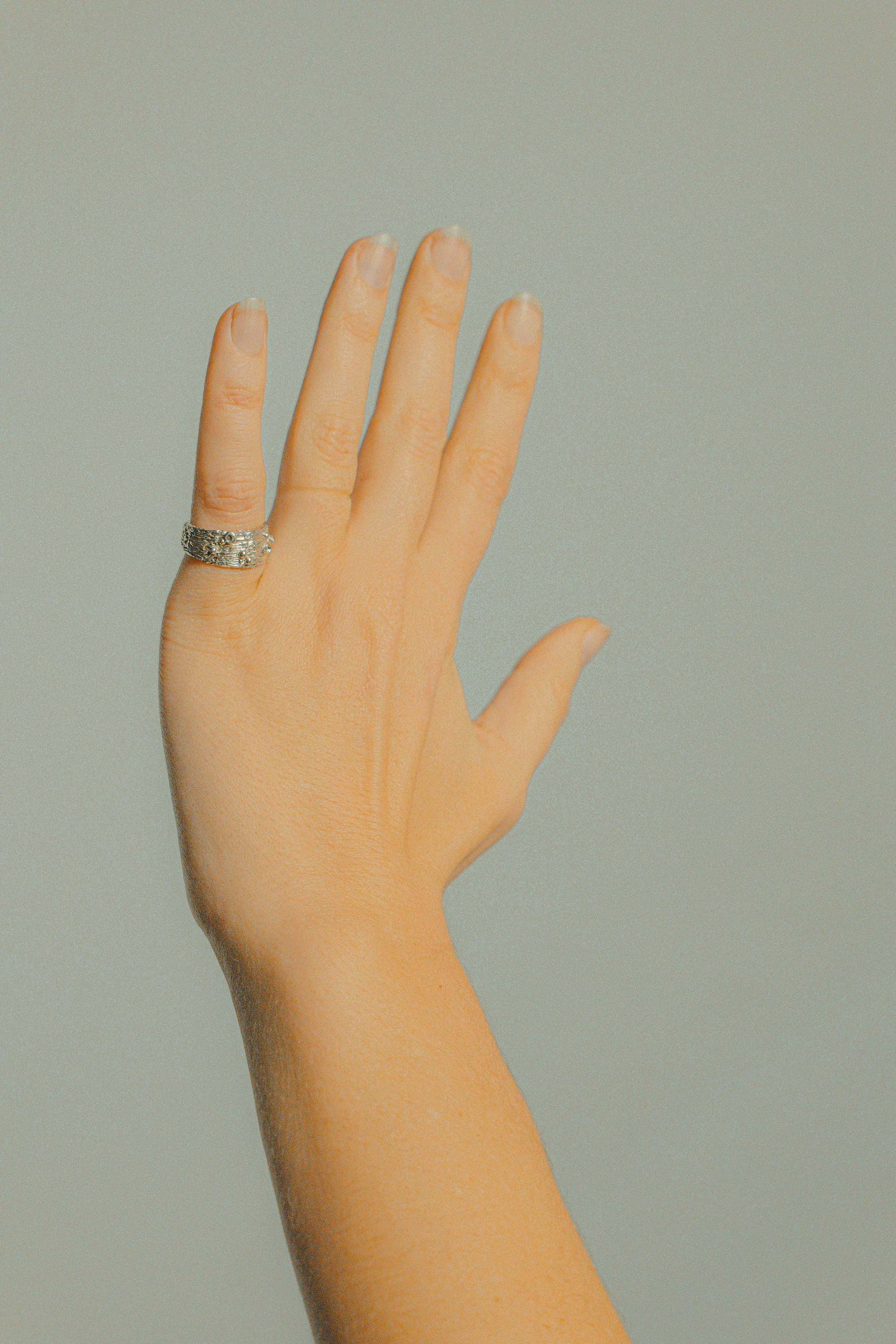 A human hand raised with fingers extended, wearing a silver ring on the index finger, against a plain background.