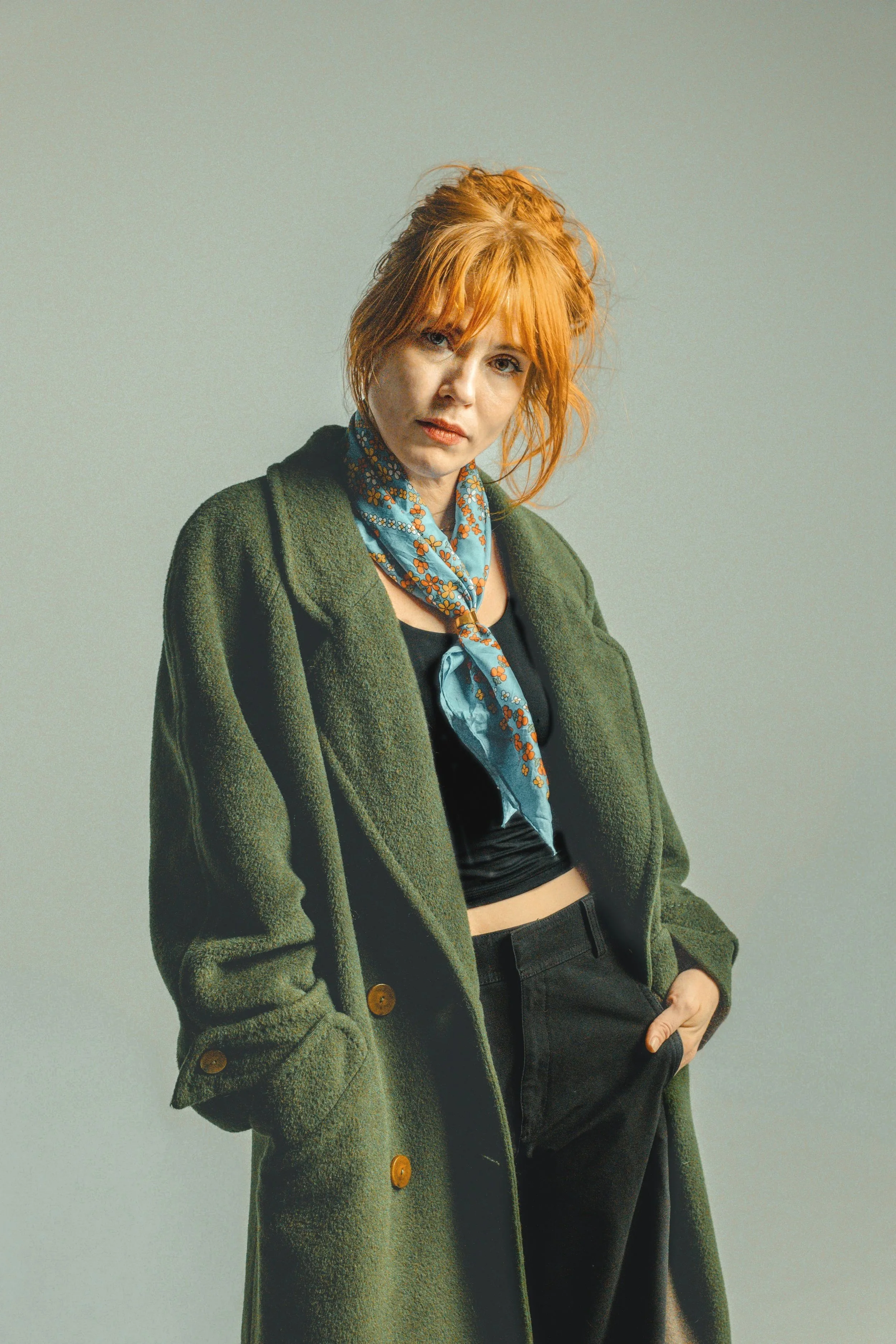A woman with red hair styled in a messy updo, wearing a green coat, black top, and a blue patterned scarf, standing against a plain background.