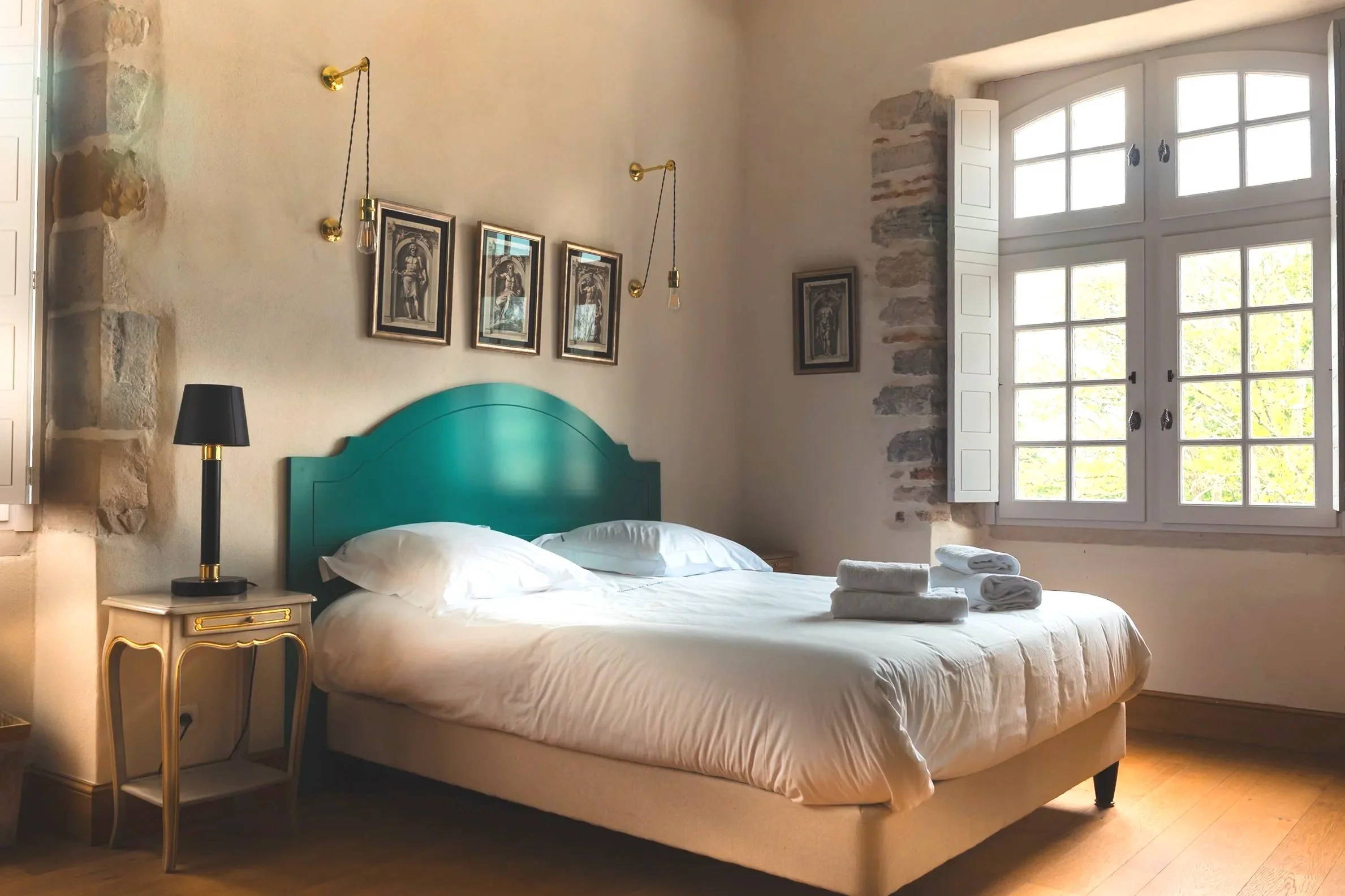 A cozy bedroom with a large window, a teal headboard, white bedding, towels on the bed, a black lamp on a vintage nightstand, and framed artwork on the wall.