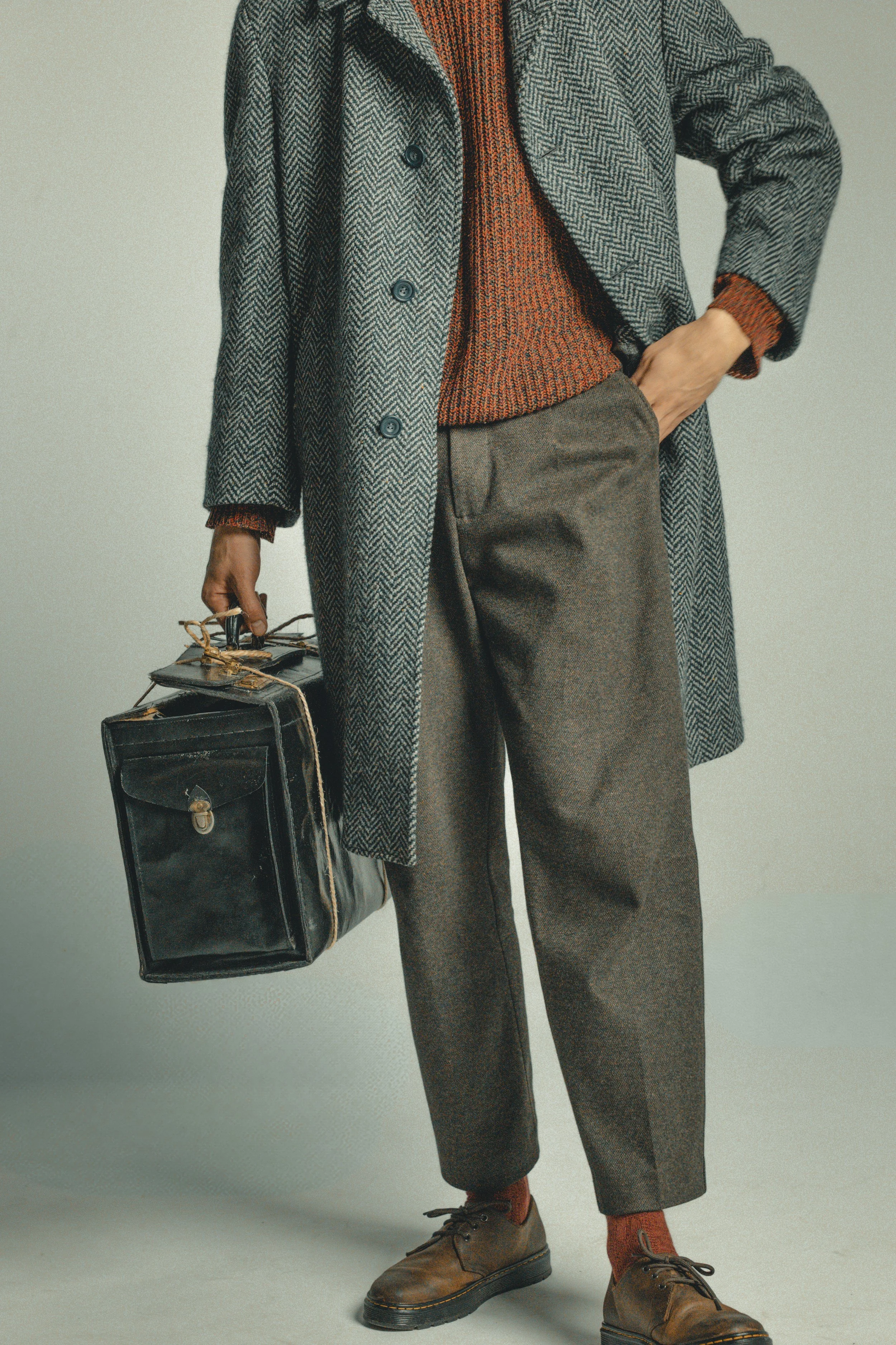 A person dressed in vintage style clothing, holding a black leather briefcase, wearing a herringbone overcoat, a rust-colored sweater, gray pants, rust-colored socks, and brown shoes.