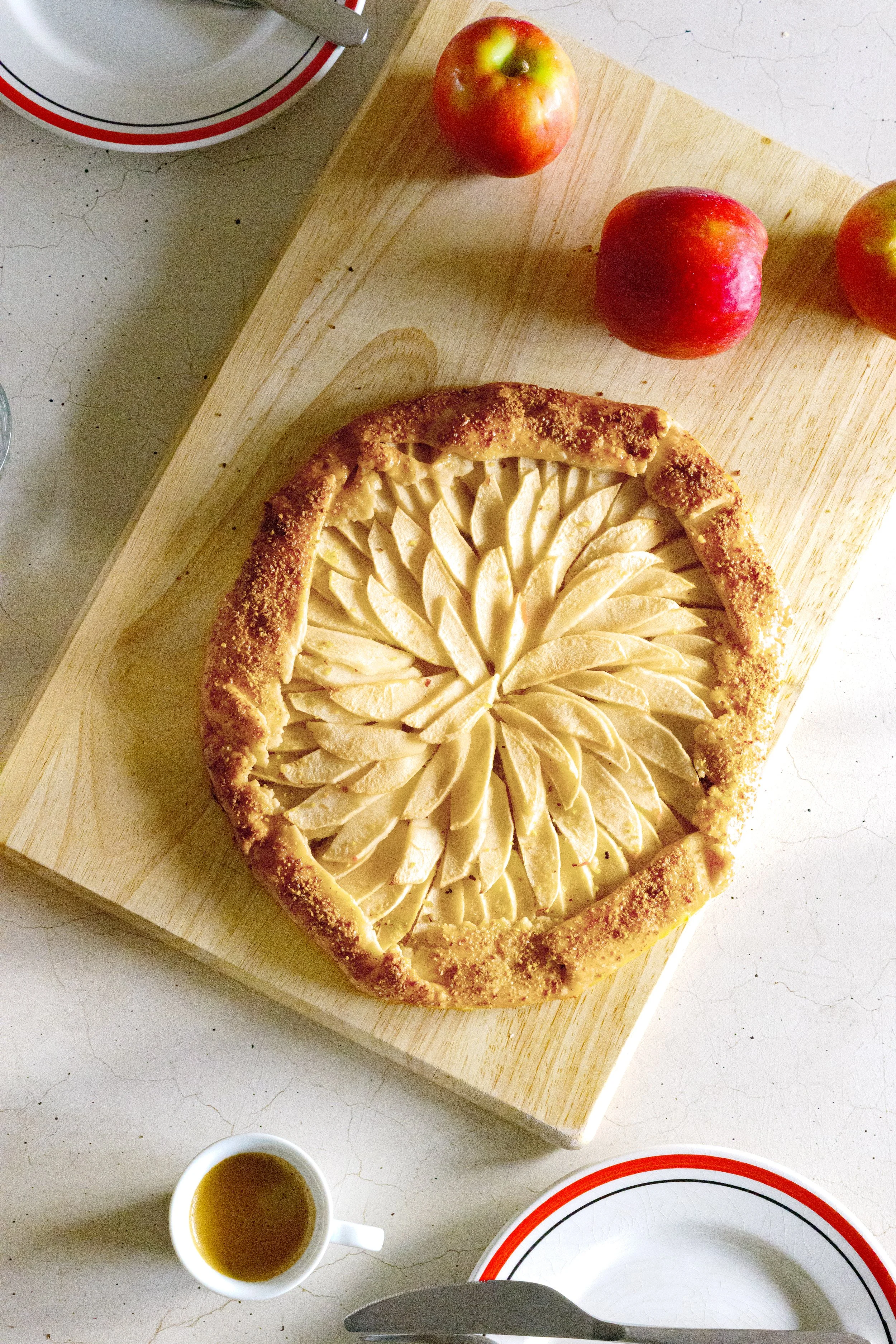 An apple tart with sliced apples arranged in a decorative pattern on a wooden board, surrounded by whole apples, a cup of tea, and plates.