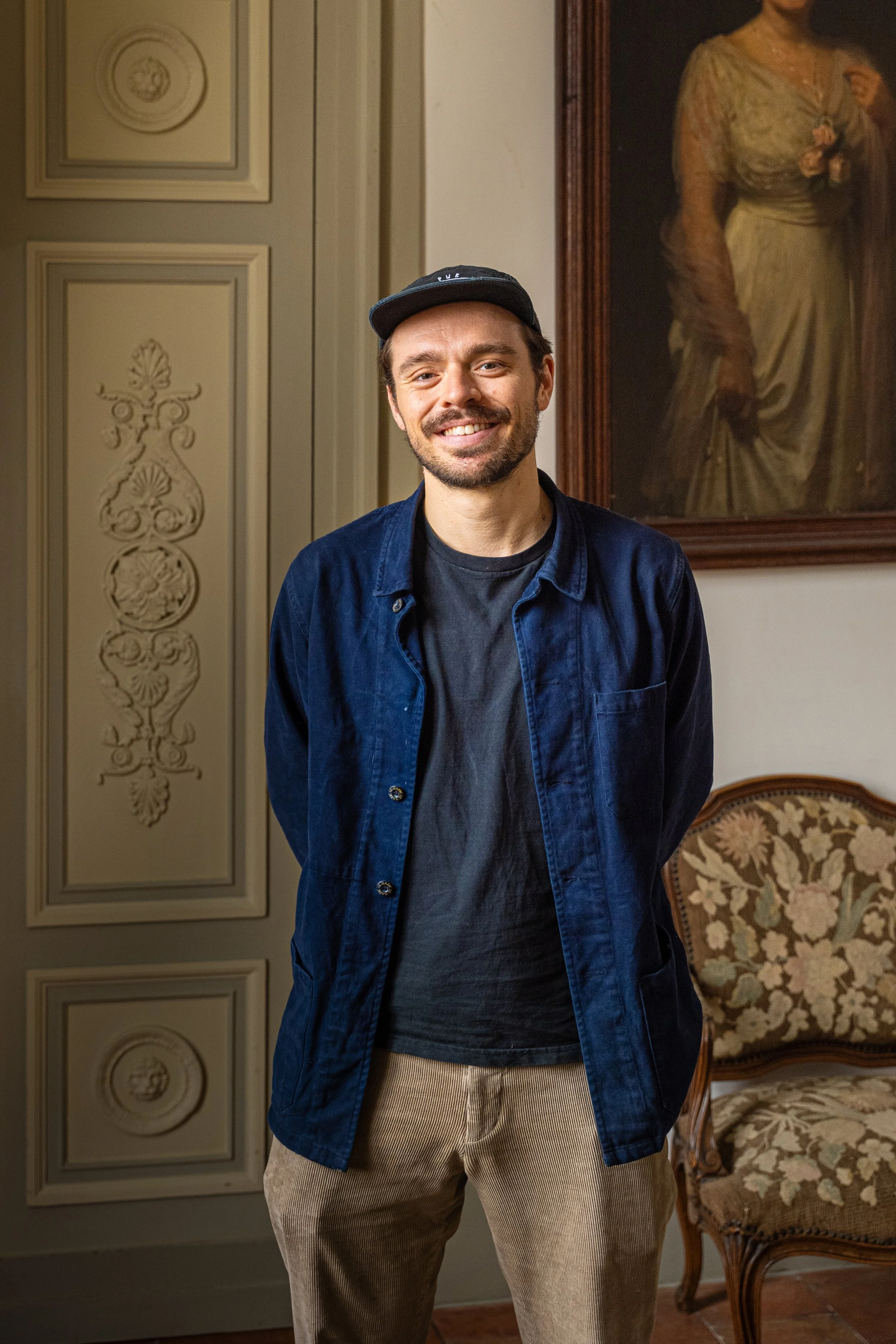 A smiling man with a beard and mustache wearing a black cap, navy blue jacket, black t-shirt, and beige pants, standing indoors in front of decorative wall panels, a framed portrait painting, and a floral-patterned antique chair.