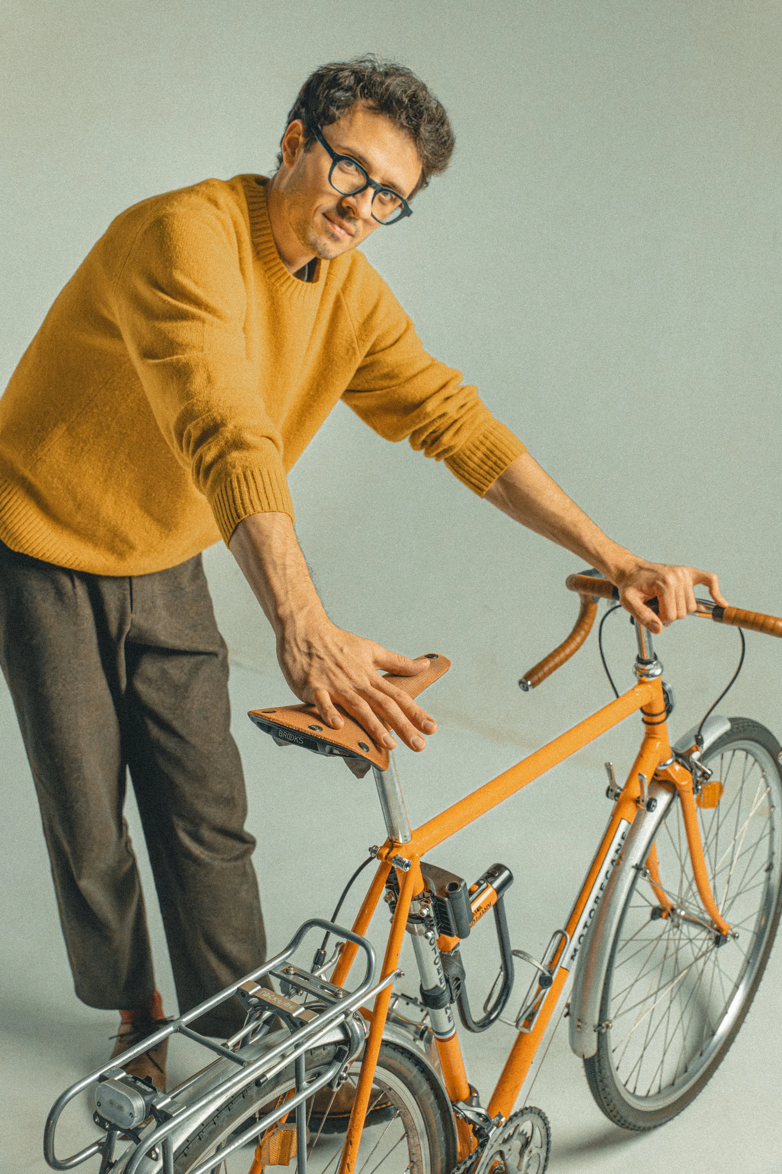 A man wearing glasses and a mustard yellow sweater, leaning on an orange bicycle with a brown saddle, against a plain background.