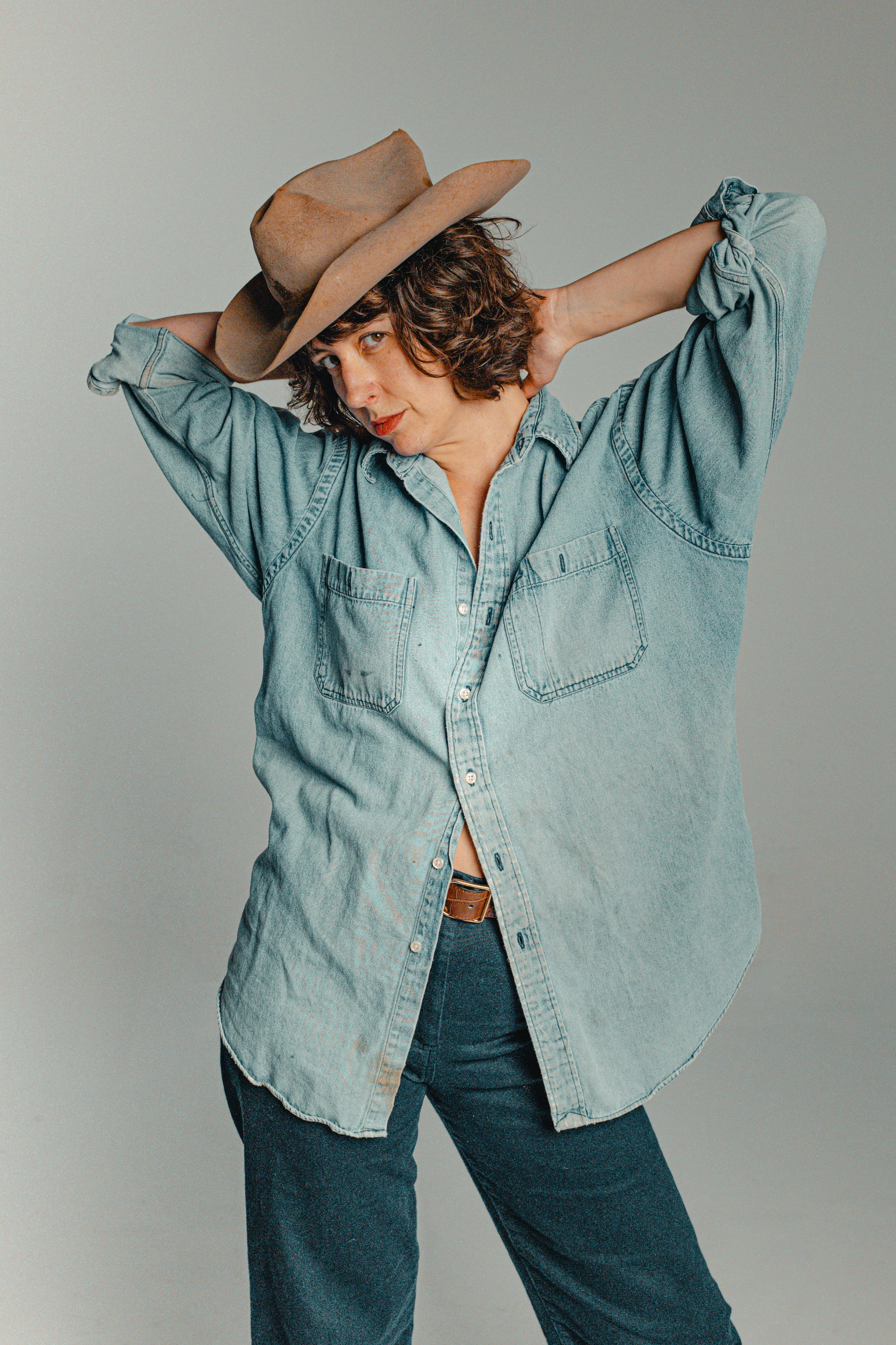 Woman in a denim shirt and jeans with a wide-brimmed hat, posing with her hands behind her head.