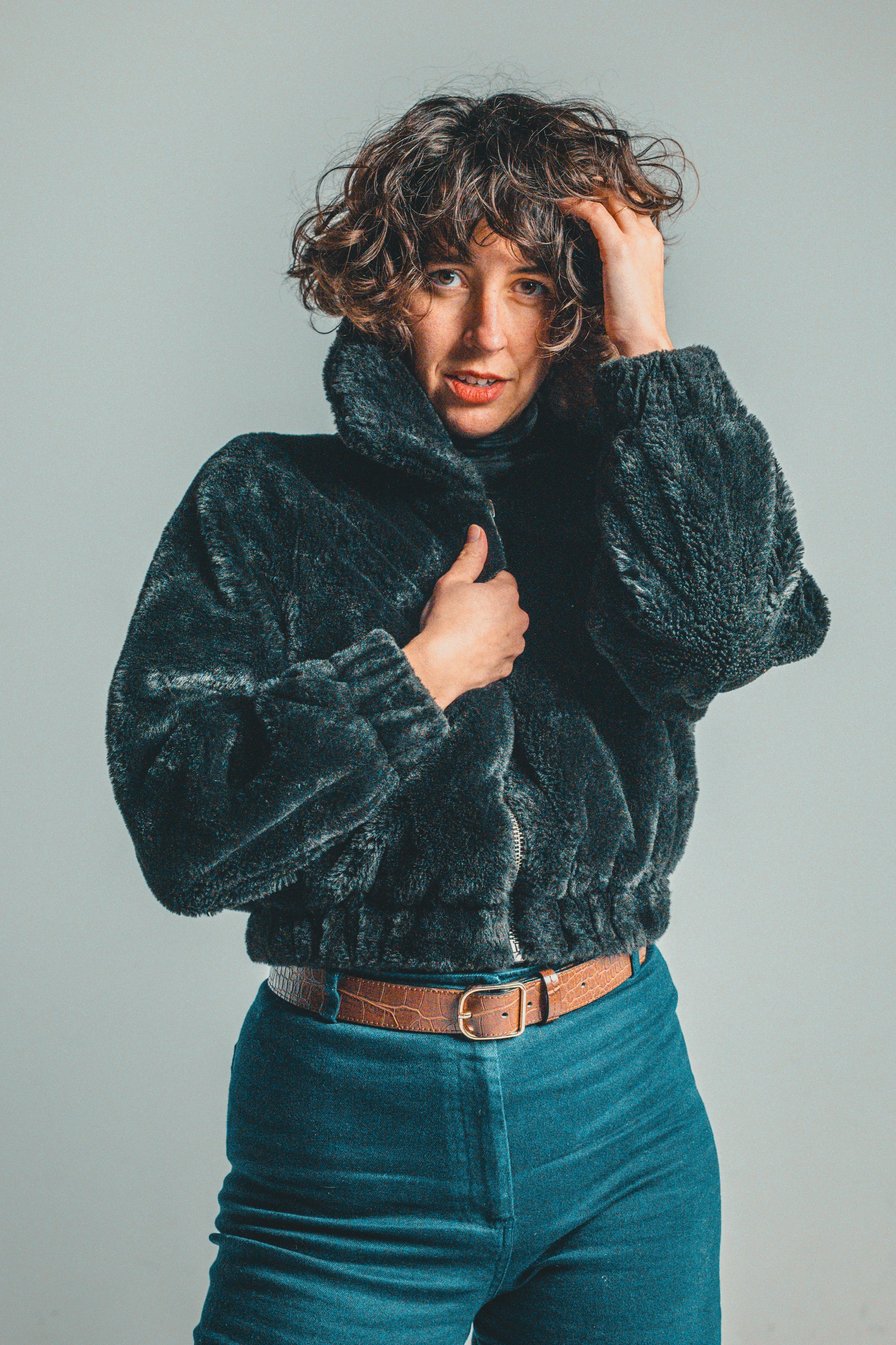 Woman with curly hair wearing a black fur jacket, standing against a plain gray background.