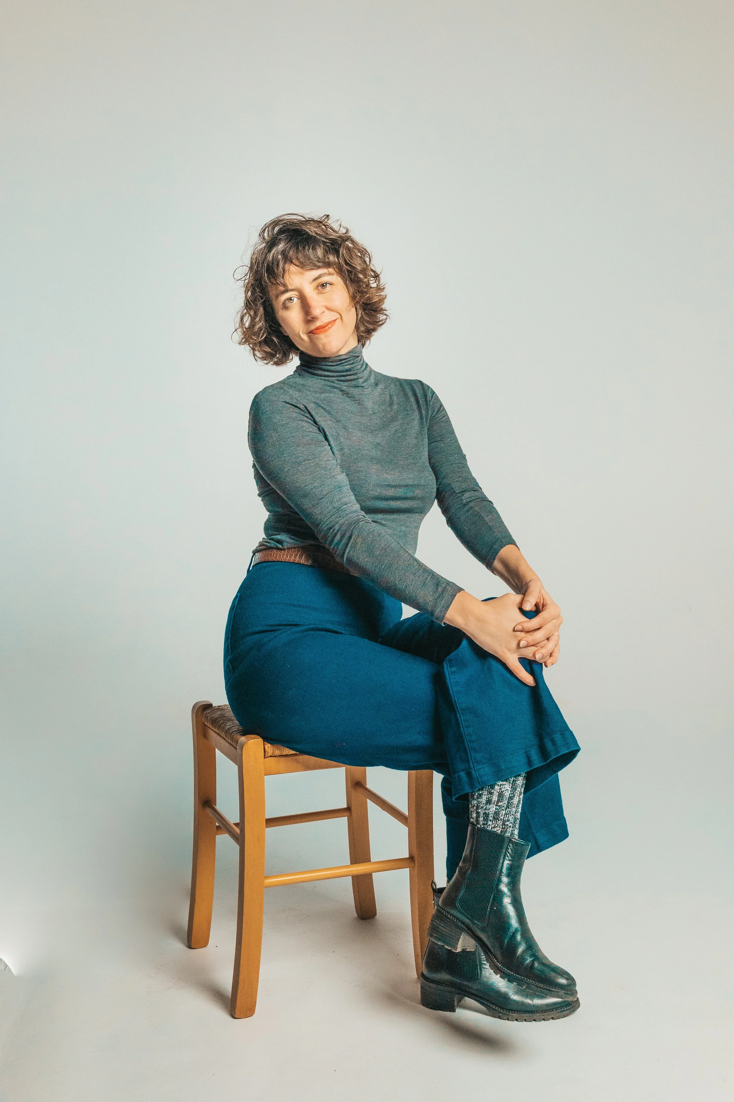 A woman with curly hair sitting on a wooden stool, wearing a gray turtleneck, blue pants, patterned socks, and black boots, posing against a light background.