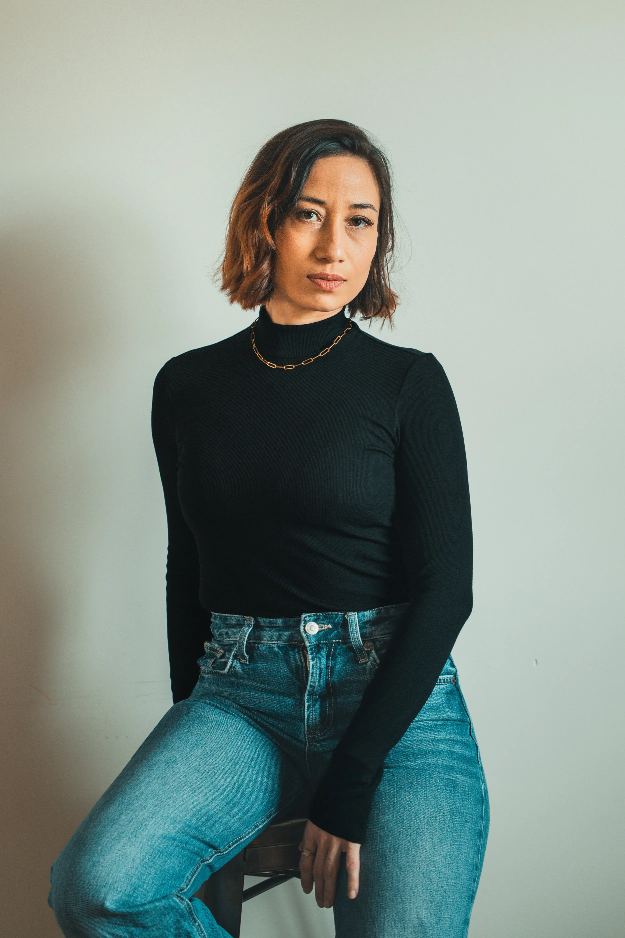 A woman with shoulder-length brown hair wearing a black turtleneck, gold chain necklace, and blue jeans, sitting on a wooden stool against a plain light-colored wall.