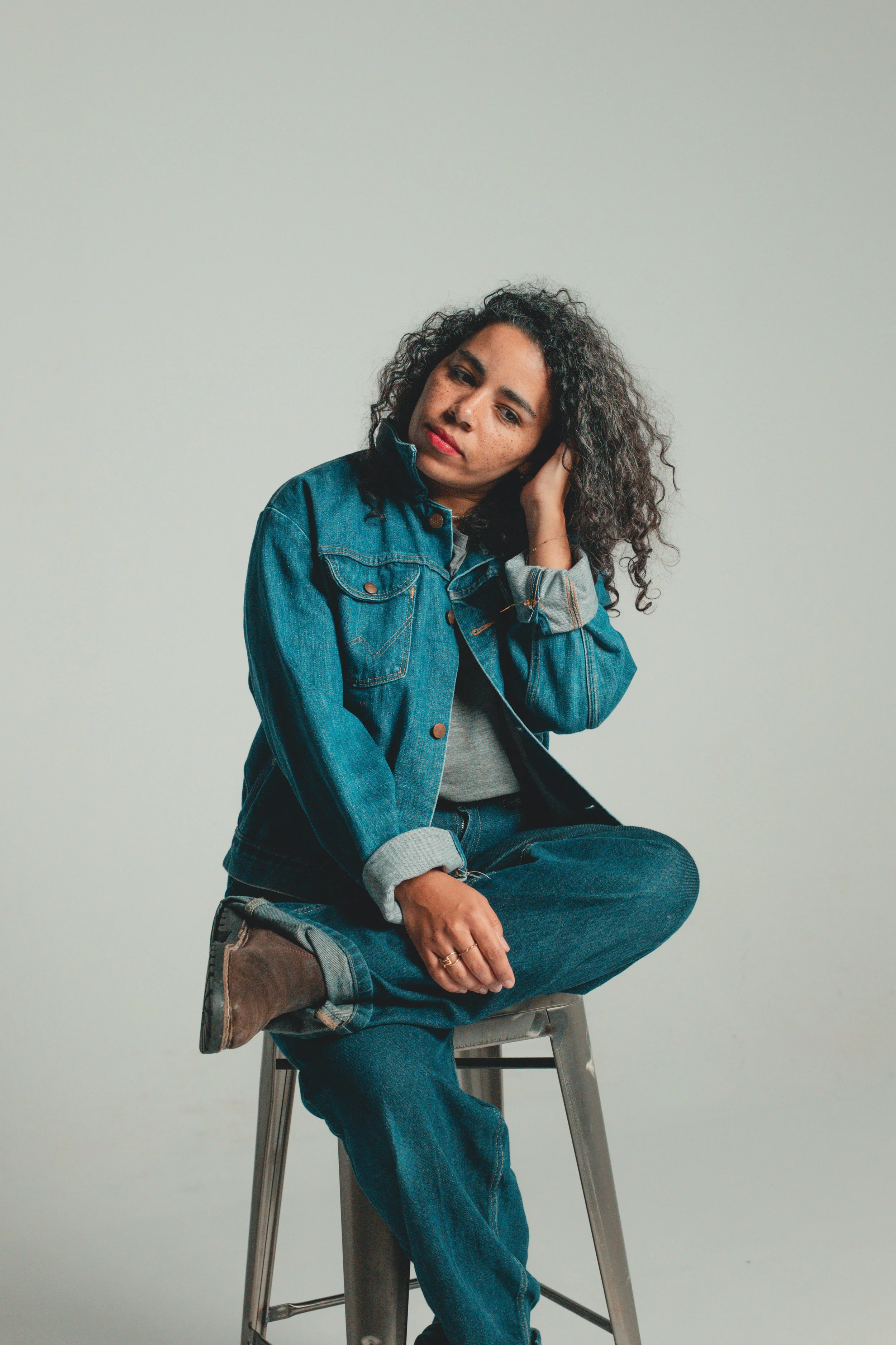 A woman with curly hair wearing a denim jacket and jeans, sitting on a metal stool with one leg crossed over the other, against a plain light background.