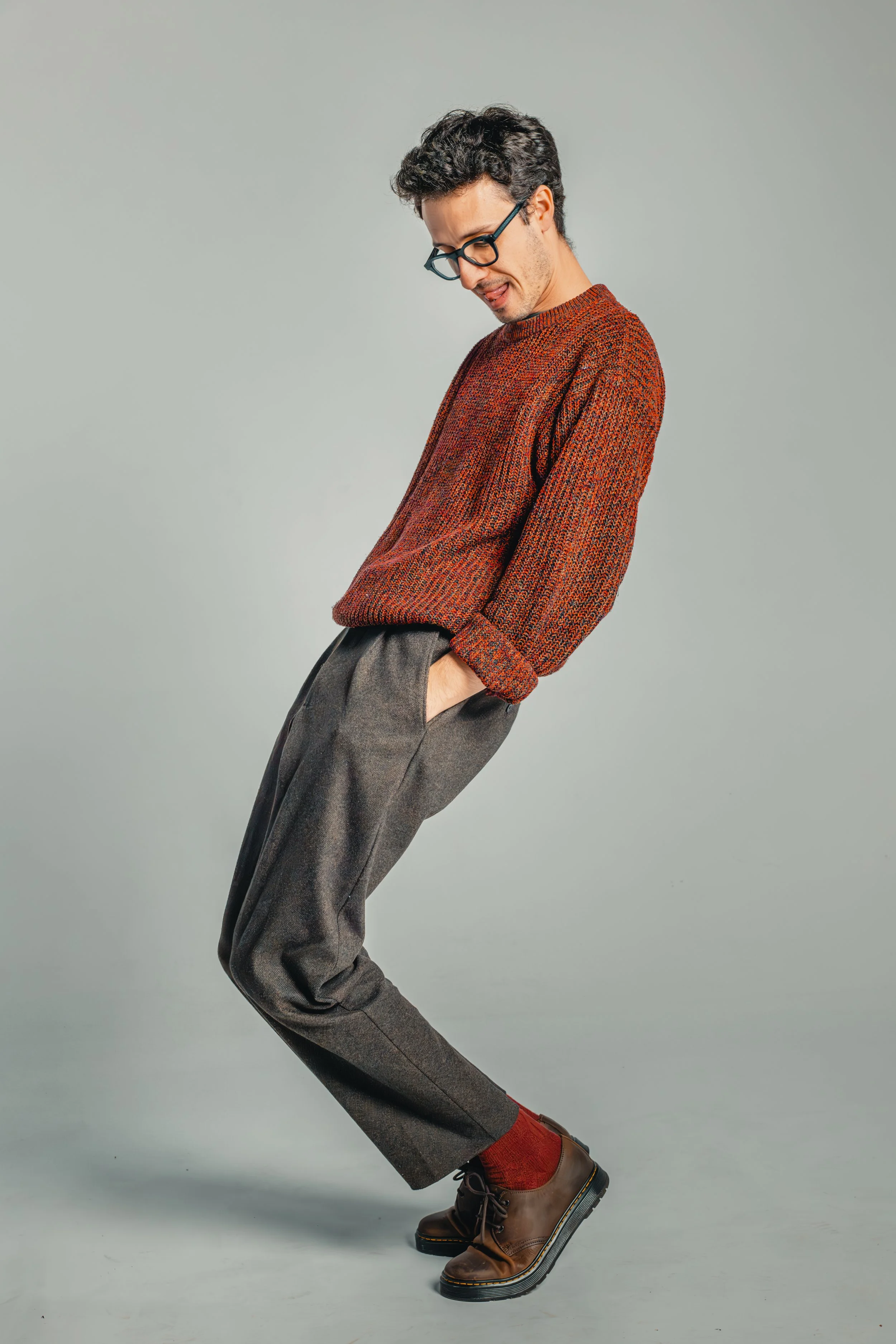A man in a rust-colored sweater, gray plaid trousers, red socks, and brown shoes is standing against a gray background, looking downward with a playful expression.