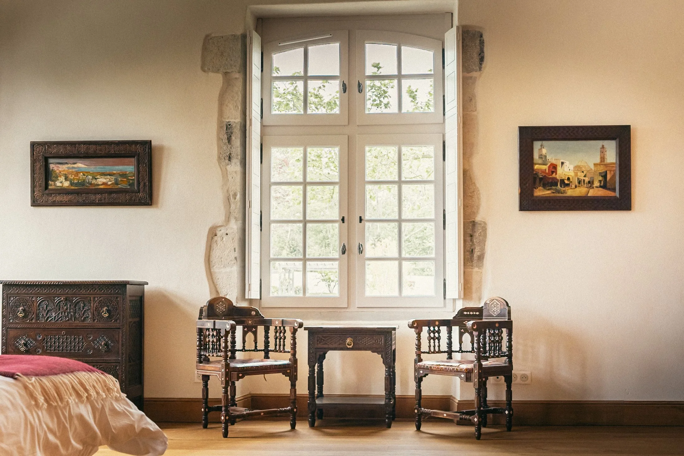 A bedroom with a large window in the center, two wooden chairs, and a matching small table in front of the window. The window has decorative shutters and reveals greenery outside. The walls are painted in a light color, with two framed paintings hang
