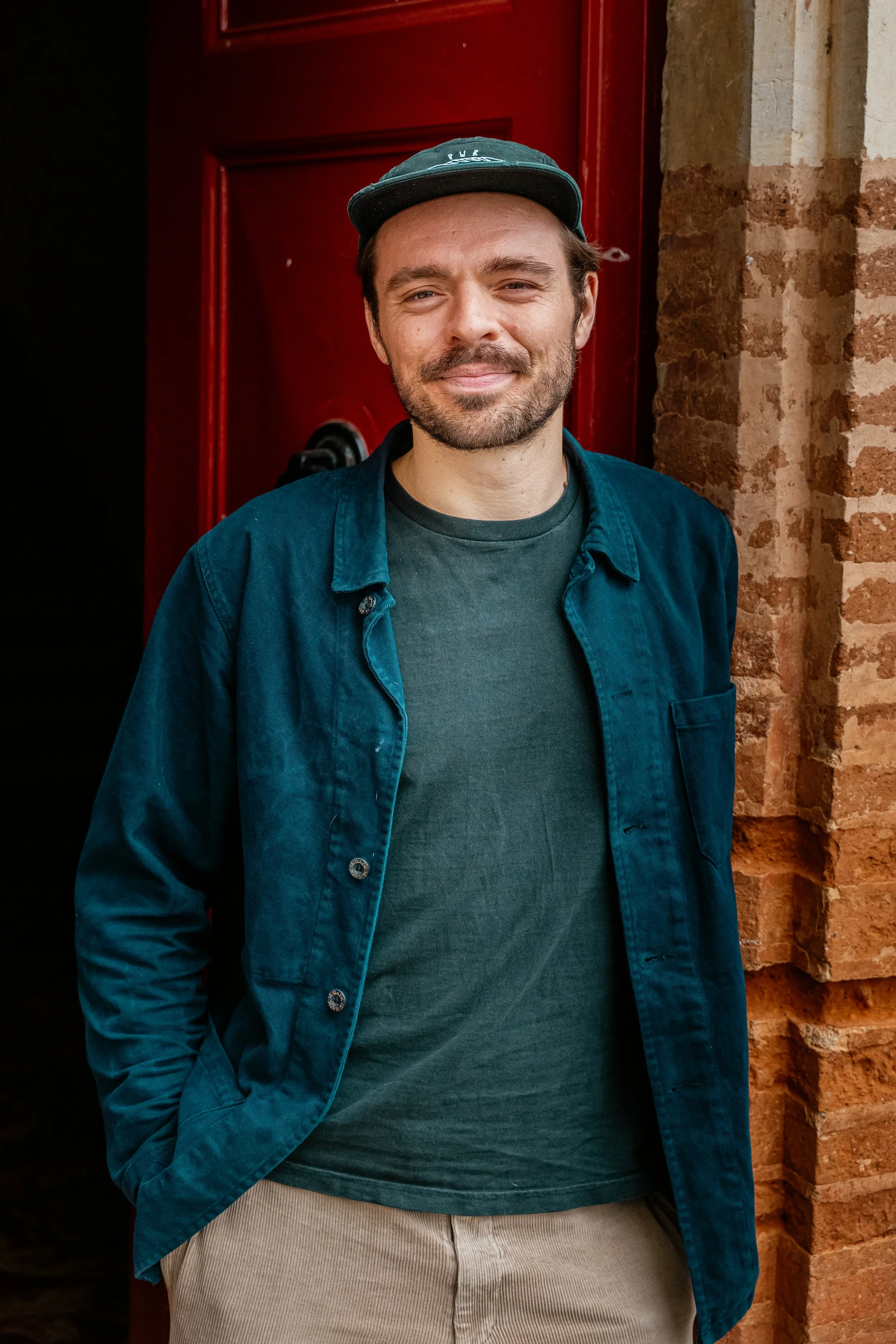 A young man in casual clothing standing outside a brick building with a red door, smiling at the camera.