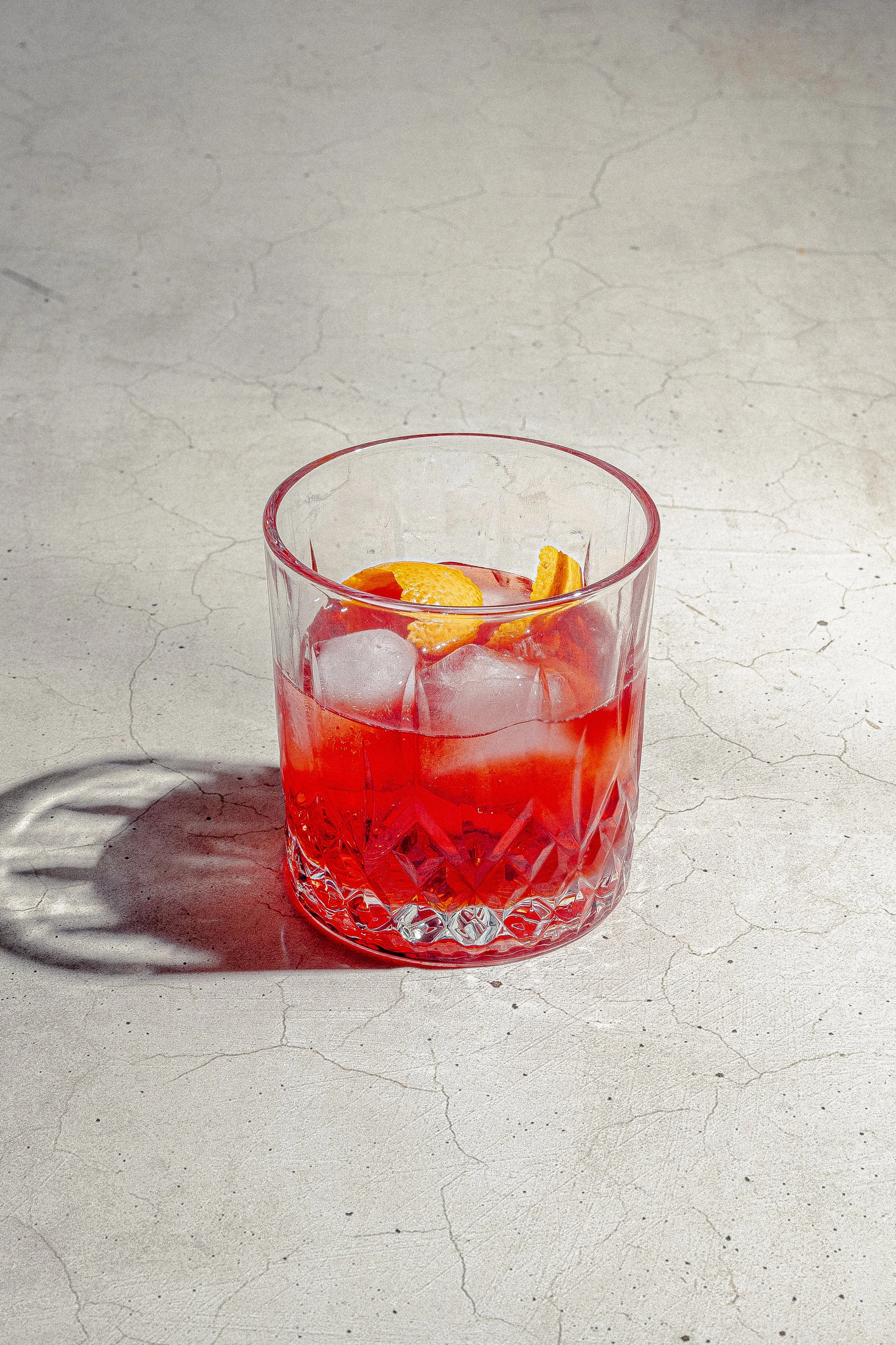 A clear textured glass tumbler filled with pink cocktail, ice cubes, and garnished with lemon peel, placed on a cracked white surface.