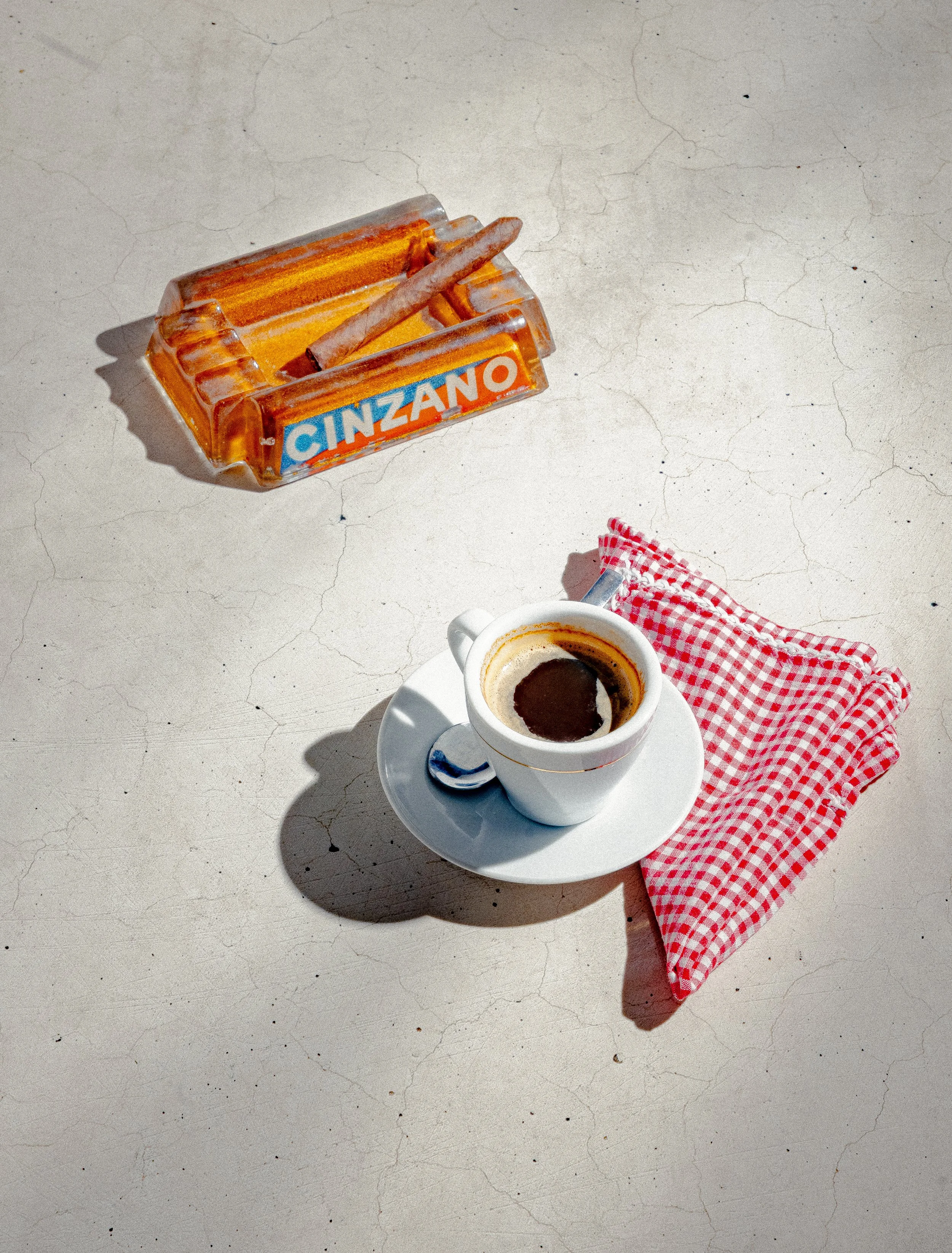 A cup of black coffee on a white saucer with a small spoon, a red and white checkered napkin, and an ashtray with a cigarette and a matchstick on a cracked white surface.