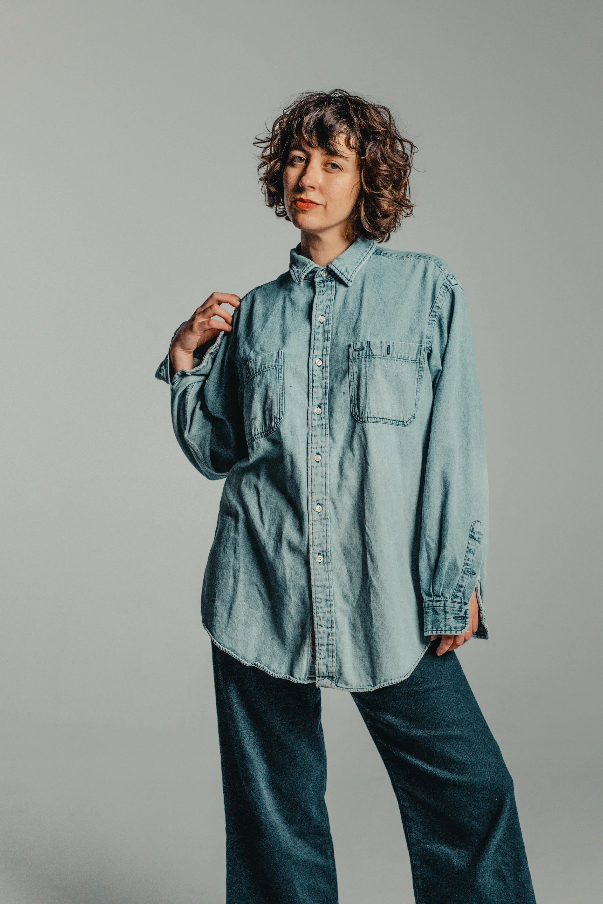 A woman with curly hair wearing a light denim shirt and dark denim pants, standing against a plain background.